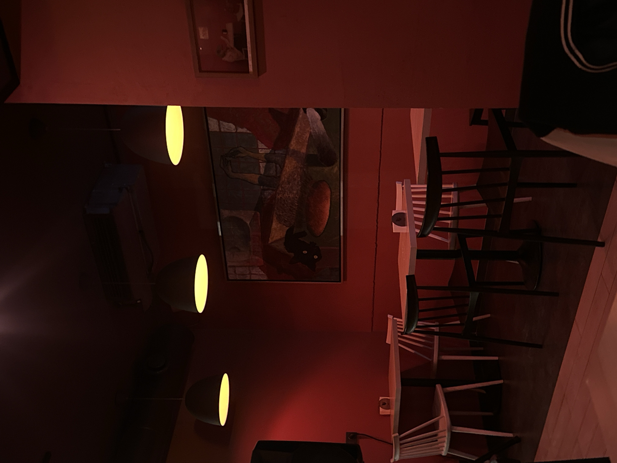 The image depicts the interior of a dimly lit space, likely a restaurant or bar, in Yerevan, Armenia. The walls are painted in a reddish hue, and several minimalist light fixtures with cylindrical shades provide illumination. Various framed artworks adorn the walls, including one with a painting of an abstract figure resembling a cat. The furniture includes a series of wooden chairs and tables, mostly in dark tones, though some chairs are painted white. A black television is placed on the wall. No people are visible in the scene.