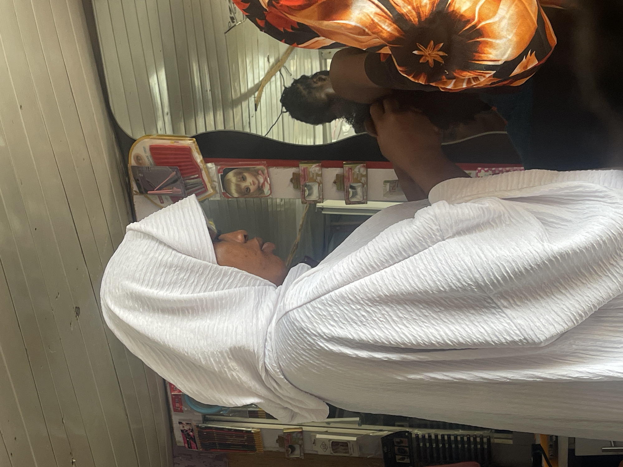 The image shows a close-up of a person from the side, wearing a white headscarf and draped in a white garment. The person appears to be having their hair styled by another individual whose hands are visible, wearing a colorful headscarf with an orange and black floral pattern. The setting appears to be a hair salon, with shelves of products and a mirror visible in the background. A doll's face is displayed on one of the shelves. The walls are made of wood panels, and the lighting suggests an indoor environment. This scene is likely taking place in Ado, Nigeria, a city known for its bustling hair salons.