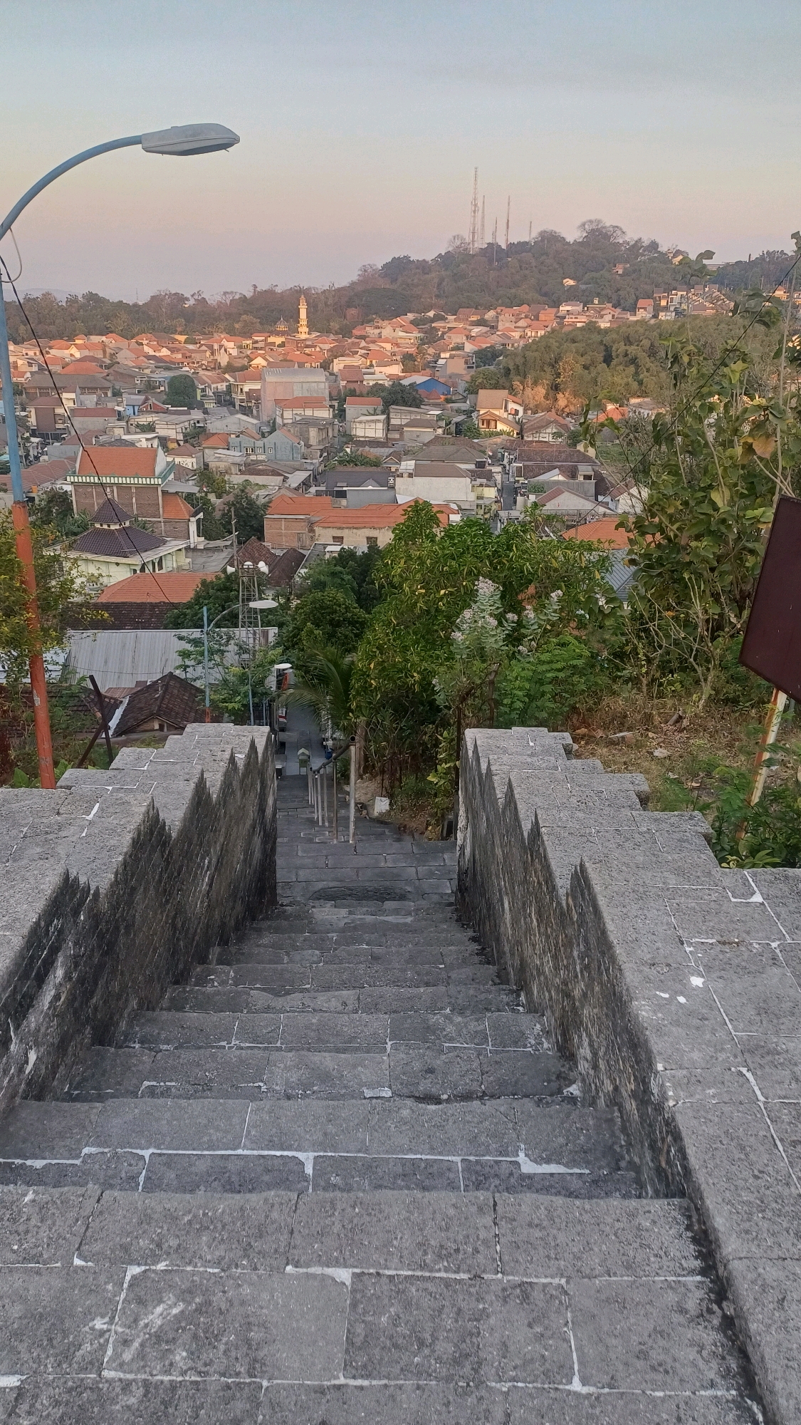 The image shows a view from a stone staircase looking down onto a town in Tlogopatut, Indonesia. The sky is clear and hazy, with a soft gradient of color indicating either sunrise or sunset. A streetlamp is visible on the left side of the image, curving into the frame. The staircase itself is made of large, gray stone blocks and is flanked by stone walls with a pointed, jagged top edge.
The town below is densely packed with buildings, many of which have orange or red tile roofs. A mosque with a tall minaret is visible in the mid-ground, and further in the distance, several radio towers stand on a hilltop. Lush green trees are interspersed throughout the town and on the surrounding hills. A narrow road runs through the town, with a few vehicles visible. The overall impression is one of a residential area nestled within a hilly landscape.