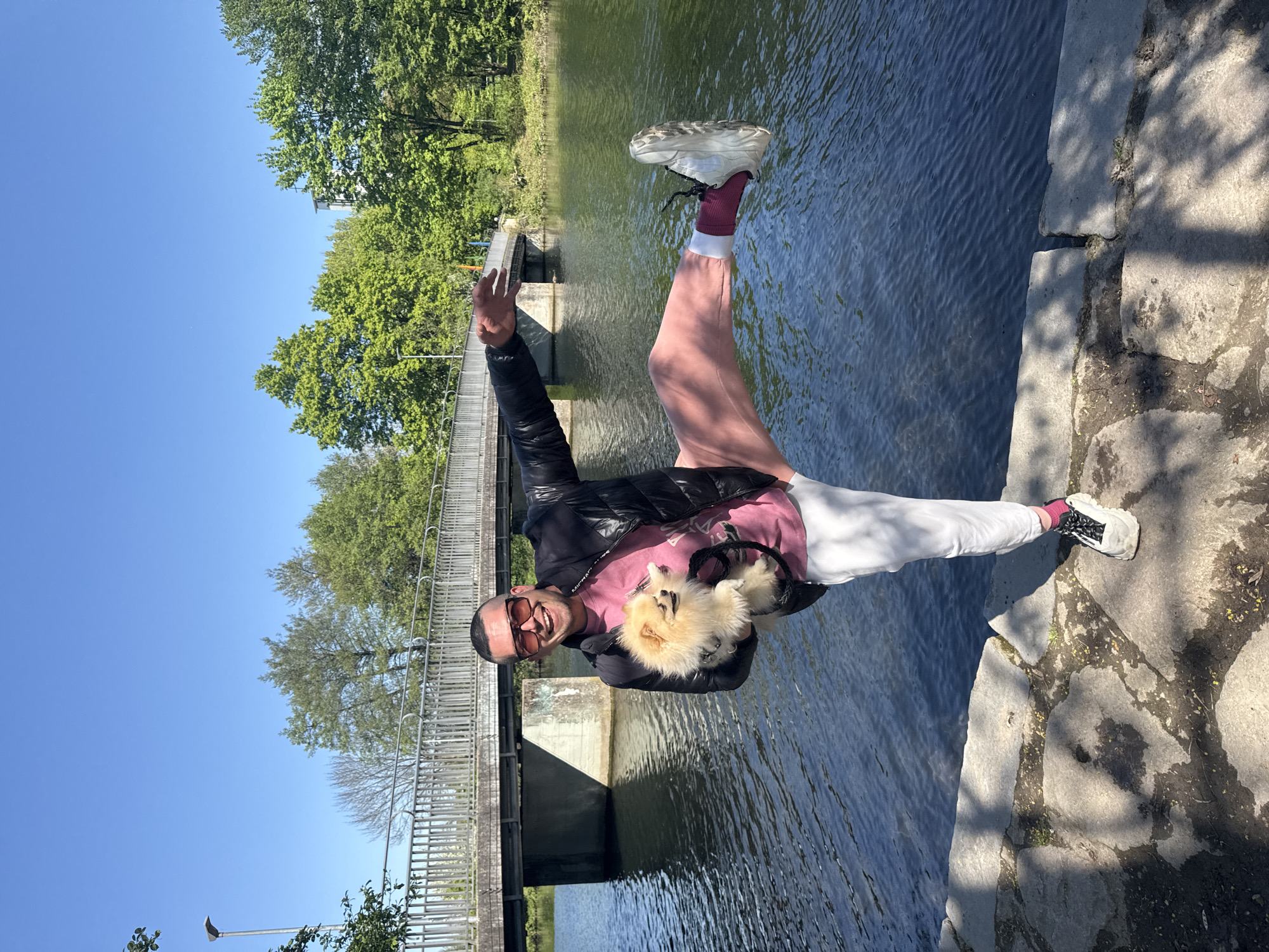 An individual is captured in a vibrant, outdoor scene in Bietigheim-Bissingen, Germany, posing energetically on the bank of a body of water, likely a river or canal. The person, a smiling man, appears to be in his late 20s or 30s, wearing sunglasses, a black puffer jacket (with "MICHAEL KORS" faintly visible on the sleeve), a pink long-sleeved shirt, pink sweatpants, and white sneakers with red socks. He is holding a fluffy, light-colored Pomeranian-like dog in his left arm, possibly in a carrier or sling.
The man is actively posing for the camera, making a peace sign with his right hand and lifting his left leg, bending it at the knee. The setting is bright and sunny, indicated by a clear blue sky and visible shadows cast on the concrete and stone pathway beside the water. Lush green trees line the far bank and parts of the background. A metal bridge with a railing spans the water further back in the image. The water surface shows ripples, reflecting the blue sky and surrounding elements. Concrete structures are visible both as the bank where the person stands and as parts of the bridge and possibly a weir in the distance. The overall impression is one of a cheerful and active outing on a pleasant day.