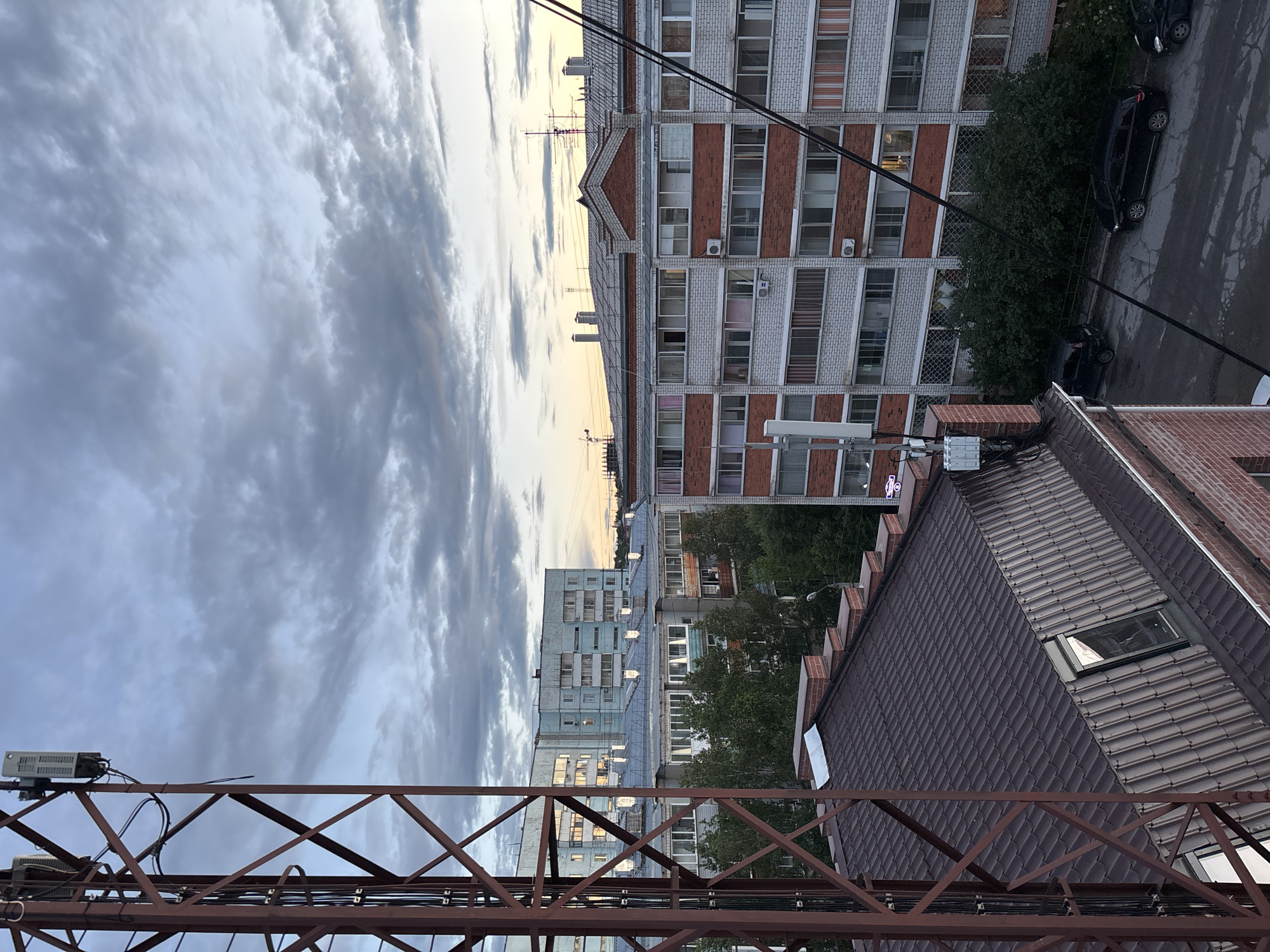 The image depicts an outdoor, elevated view of an urban residential area in Blagoveshchensk, Russia, under a cloudy sky during what appears to be twilight. In the foreground, a tall, brown metal lattice tower, possibly a telecommunications mast, dominates the left side of the frame. Several white electronic devices are mounted on the upper section of the tower.
Behind the tower, a multi-story brick building with a dark brown, corrugated metal roof is visible, featuring a dormer window. Further back, a series of apartment buildings are lined up. The closest apartment building is a mid-rise structure with a facade of white brick and red brick accents. It has multiple floors with numerous windows, some with balconies, and air conditioning units are visible. Another apartment building, taller and painted a light blue, is situated further in the background, with its windows reflecting the subdued light.
The street level below the buildings shows asphalt pavement with parked black cars. Dense green trees and bushes are interspersed between the buildings and along the street. The overall impression is of a typical residential neighborhood in a Russian city during the transition from day to night, with evidence of communication infrastructure.