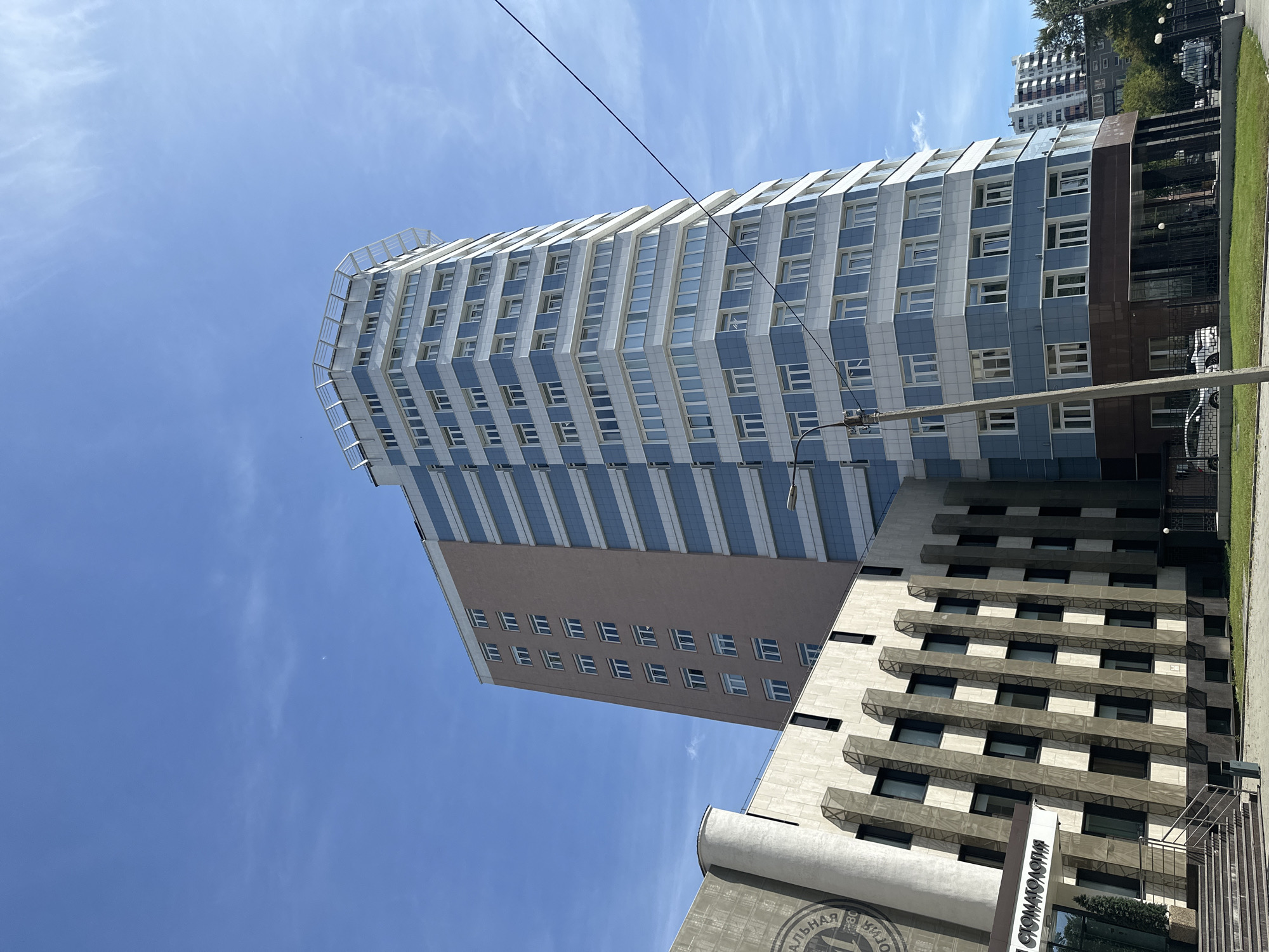 The image shows a street-level view of a modern, multi-story building with a light blue and white facade, featuring many windows. The building is tall and appears to be situated in a city. To the left of the main building, there is another, shorter building with a more textured facade in shades of beige and brown, with dark rectangular windows. This building has an entrance with stairs and a sign that reads "СТОМАТОЛОГИЯ". The sky above is clear blue with a few wispy clouds. In the background, further down the street, another residential building is visible. There are also power lines and streetlights in the foreground. The overall impression is of an urban setting on a bright day.
