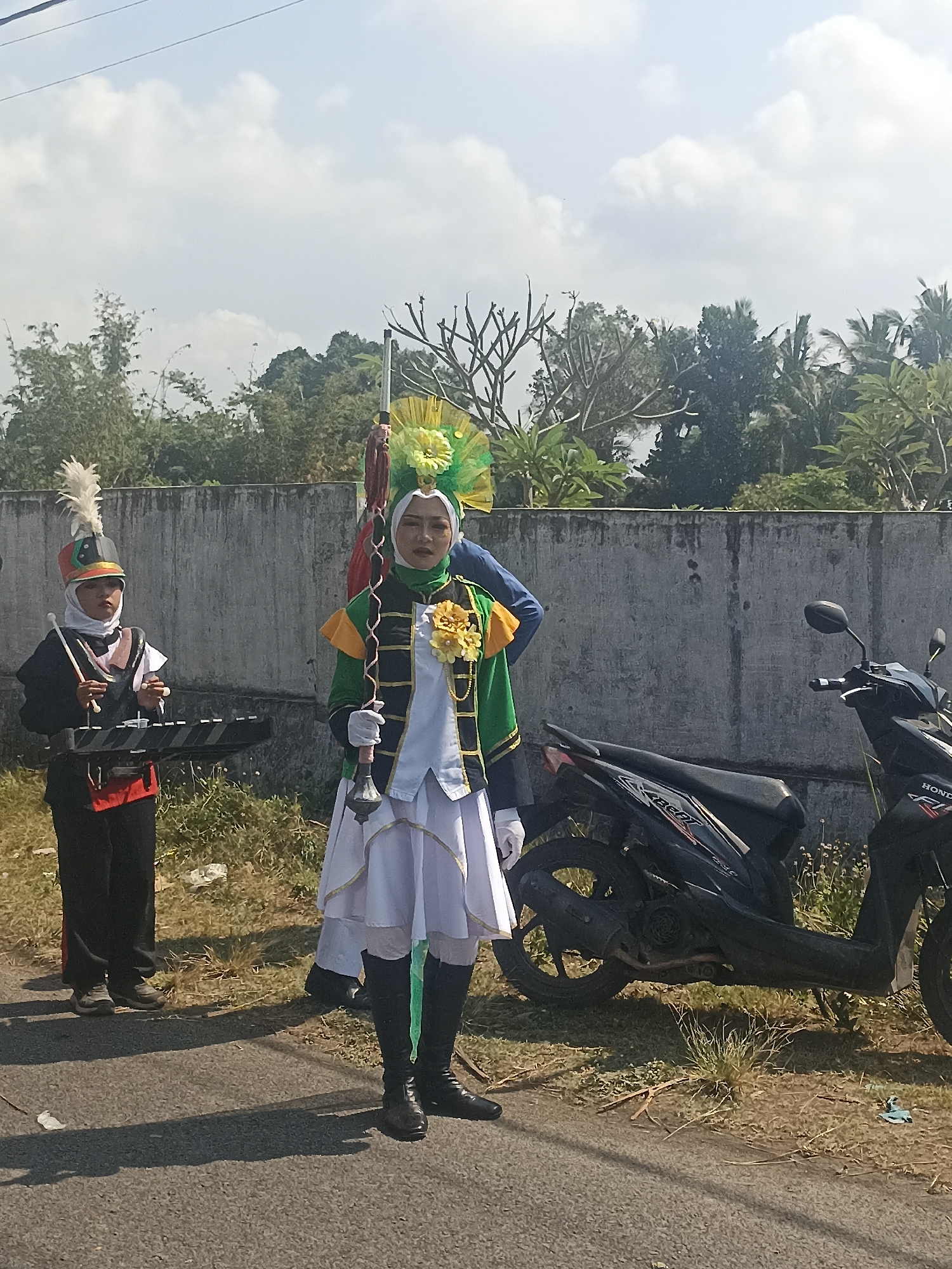 The image shows two individuals in marching band uniforms standing outdoors on a paved surface next to a concrete wall and overgrown vegetation. The person on the left wears a black jacket with red accents, a white undershirt, a red sash, and a tall hat with a white feather plume. They are holding drumsticks and a black electronic keyboard. The person on the right wears a green jacket with yellow and white trim, a white skirt, and black boots. They are holding a decorative staff. A motorcycle is parked to the right of the individuals. The sky is visible in the background with scattered clouds, and trees and foliage are present behind the wall. The scene suggests a performance or practice session for a marching band, possibly in a public area. The location is identified as Banyuwangi, Indonesia.