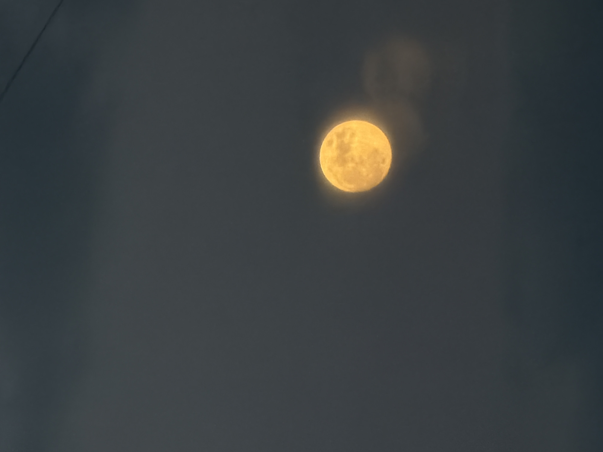 The image captures a full, luminous yellow-orange moon set against a dark, hazy sky. The moon is the central focal point, positioned slightly right of the center of the frame, emitting a soft, diffused glow. Its surface, while generally appearing smooth due to the atmospheric conditions, shows subtle textural variations. Above and to the left of the moon, two indistinct, blurry circular shapes, possibly lens flares or reflections, are visible against the dark sky. A thin, dark diagonal line, which appears to be a wire or power line, crosses the upper left corner of the image. The sky itself is a deep, uniform grey-blue, suggesting a nighttime scene with significant cloud cover or atmospheric haze, which contributes to the soft, ethereal appearance of the moon. No discernible activity is taking place other than the observation of the moon. The setting is likely outdoors at night, possibly in Michurinsk, Russia, given the provided context. No text is visible in the media file.
