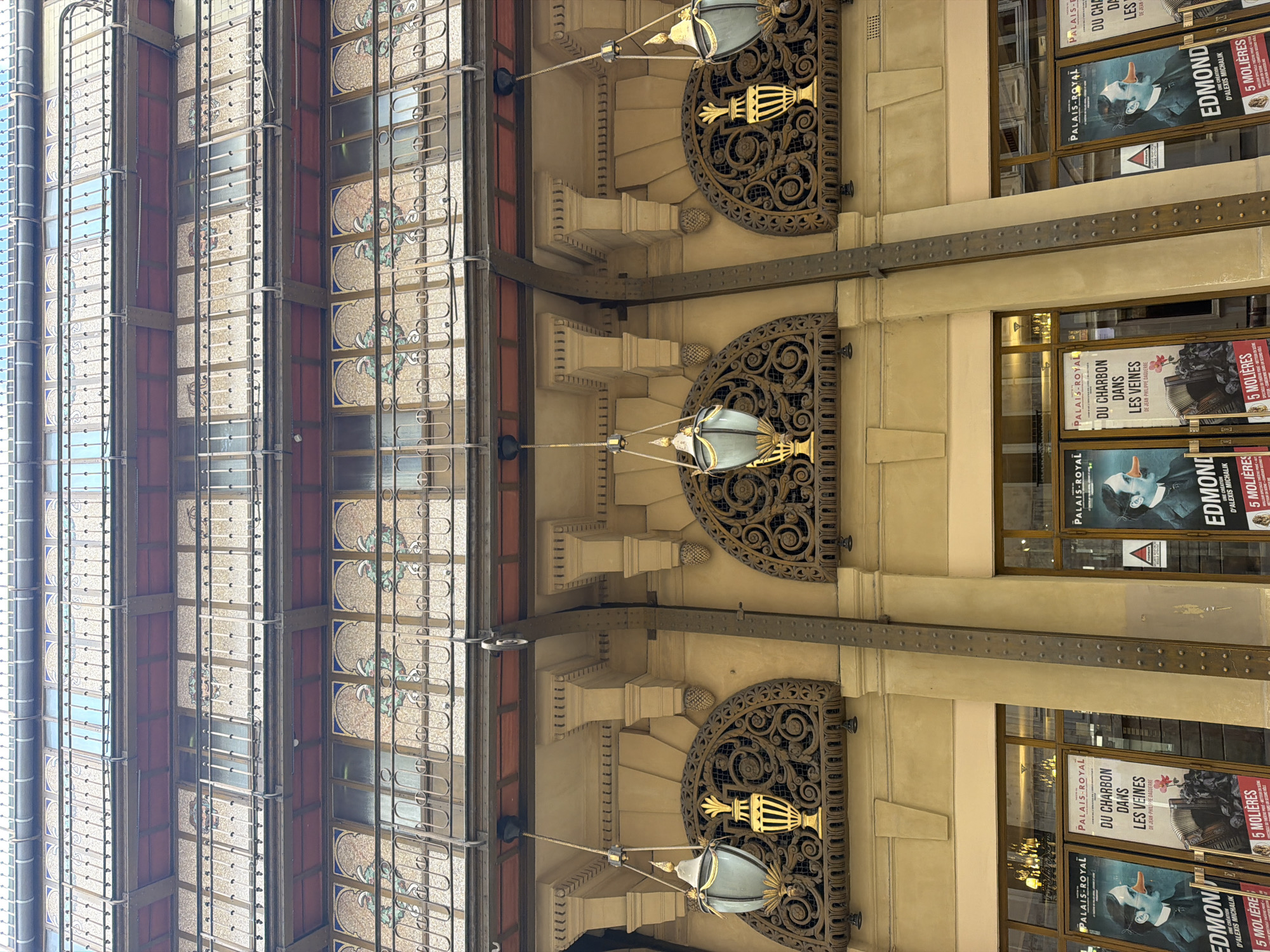 The image presents an upward view of the ornate facade of the Théâtre du Palais-Royal, situated within the Palais-Royal complex in Paris, France. The building's structure reveals multiple stories characterized by a blend of decorative and functional elements.
The upper visible levels feature balconies with white, intricate metal railings composed of repeating circular and square patterns, set against a background of mosaic-like panels displaying blue and green organic designs behind the windows. These levels are supported by visible dark metal girders, indicating a robust internal framework.
Below these, a prominent architectural band in beige houses three distinct arched recesses, each filled with dark, intricately scrolled metal grilles. Suspended from the soffit of this section are three decorative light fixtures, each comprising a light blue-grey glass housing and a gold-colored, urn-shaped base. Vertical, dark, riveted metal beams are integrated into the facade, providing structural support from the upper levels down to the ground floor.
The ground level features large display windows or doors. Within these windows, multiple theatrical posters for the "PALAIS-ROYAL" are displayed. Notable advertisements include "EDMOND," featuring a black and white image of a man in a suit with an elongated orange nose, accompanied by text indicating "UNE CRÉATION D'ALEXIS MICHALIK" and "5 MOLIERES." Another poster advertises "DU CHARBON DANS LES VEINES," depicting an accordion and a red flower. Reflections of interior light fixtures, possibly chandeliers, are visible in the glass of the ground-level windows. No individuals are visible in the scene.