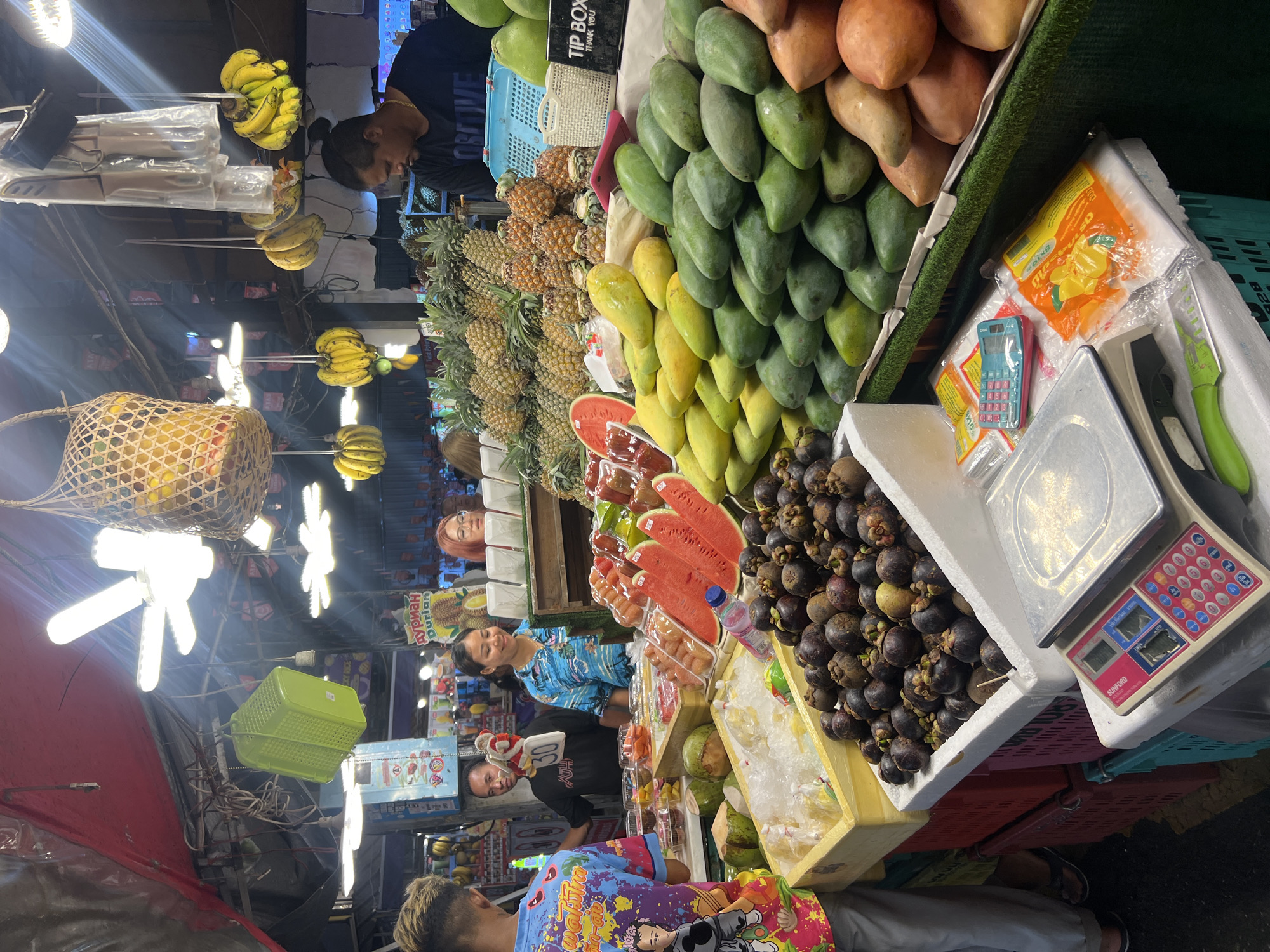 This media file captures a vibrant night market scene in Karon, Thailand, likely in the evening or at night, given the artificial lighting. The primary subjects are a variety of fresh fruits displayed for sale, including bananas, pineapples, mangoes, watermelons, and mangosteens. Several people are present, appearing to be vendors and customers interacting at the stalls.
The scene is set at a market stall overflowing with colorful produce. Objects of note include a woven basket filled with fruit, multiple green baskets, a digital scale with a calculator, and various packaging materials. The lighting appears to be from overhead fluorescent bulbs, contributing to a bustling atmosphere.
One person, a woman in a blue and white patterned top, is visible behind a fruit stall, possibly serving a customer. Another person, a man in a black t-shirt, is also visible. The overall impression is one of an active and lively market environment, with an emphasis on the abundance and freshness of the fruit offerings. There is no clear indication of the weather, but the indoor or covered nature of some stalls suggests it could be a humid tropical evening.
Visible text includes "TIP BOX THANK YOU" on a sign near the top of the frame. Another sign reads "DURIAN" in Cyrillic script, indicating that durian fruit is also sold at this market. The overall mood is one of commerce and community, with the vibrant colors of the fruit adding to the visual appeal.