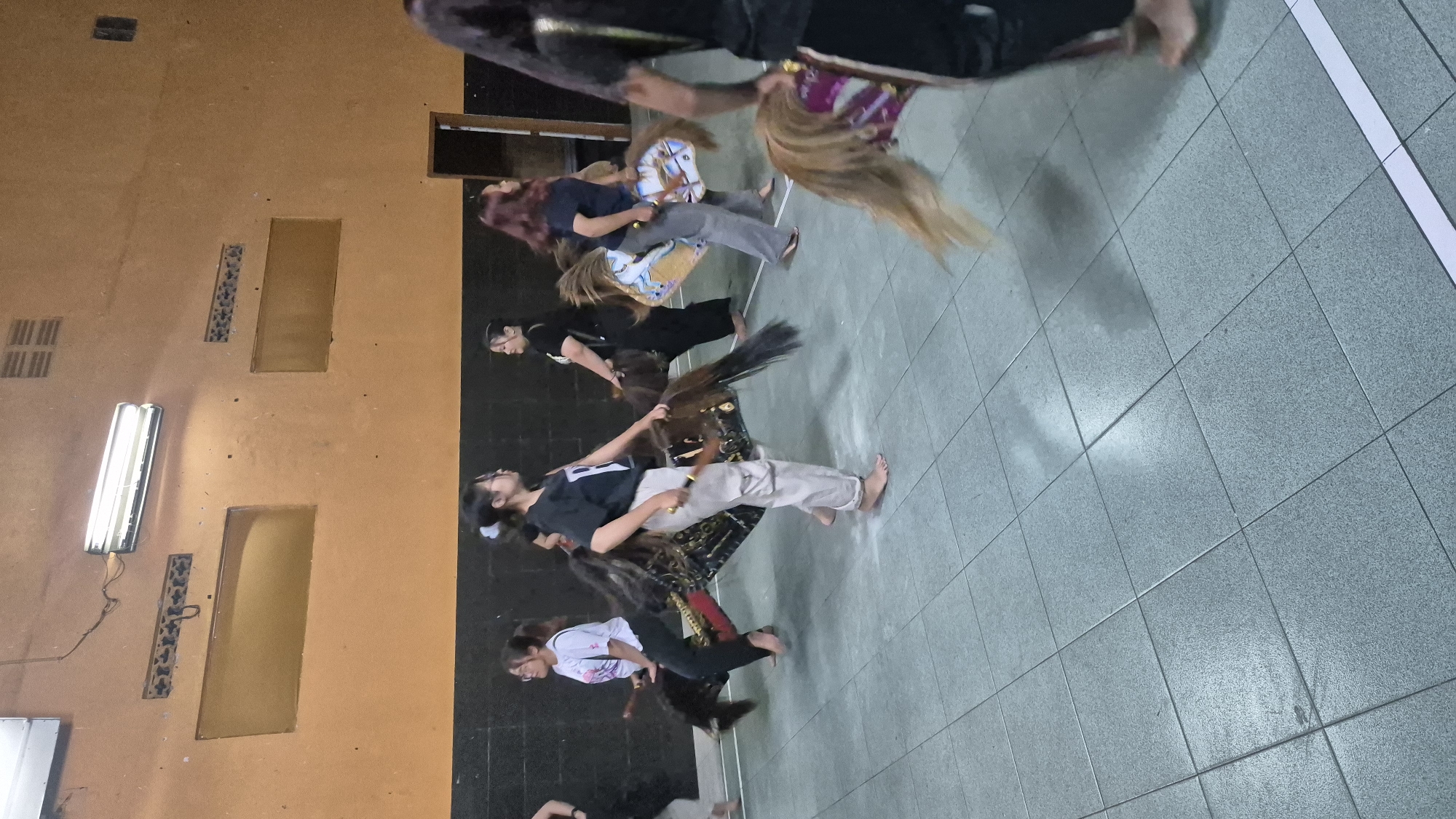 In Sumowono, Indonesia, a group of young women are practicing a traditional dance performance indoors. They are dressed in casual attire, and some are holding wooden sticks. Each performer is positioned behind a large, decorative horse puppet, which appears to be made of fabric and has a horse-like mane and tail. The puppets are supported by poles, and the women are either seated or crouched behind them, mimicking riding the horses. The setting is a large, open room with a tiled floor and an orange-painted wall. Fluorescent lights are mounted on the ceiling. The dancers are moving in unison, suggesting a rehearsal for a performance. The atmosphere is one of focused practice and cultural engagement.
