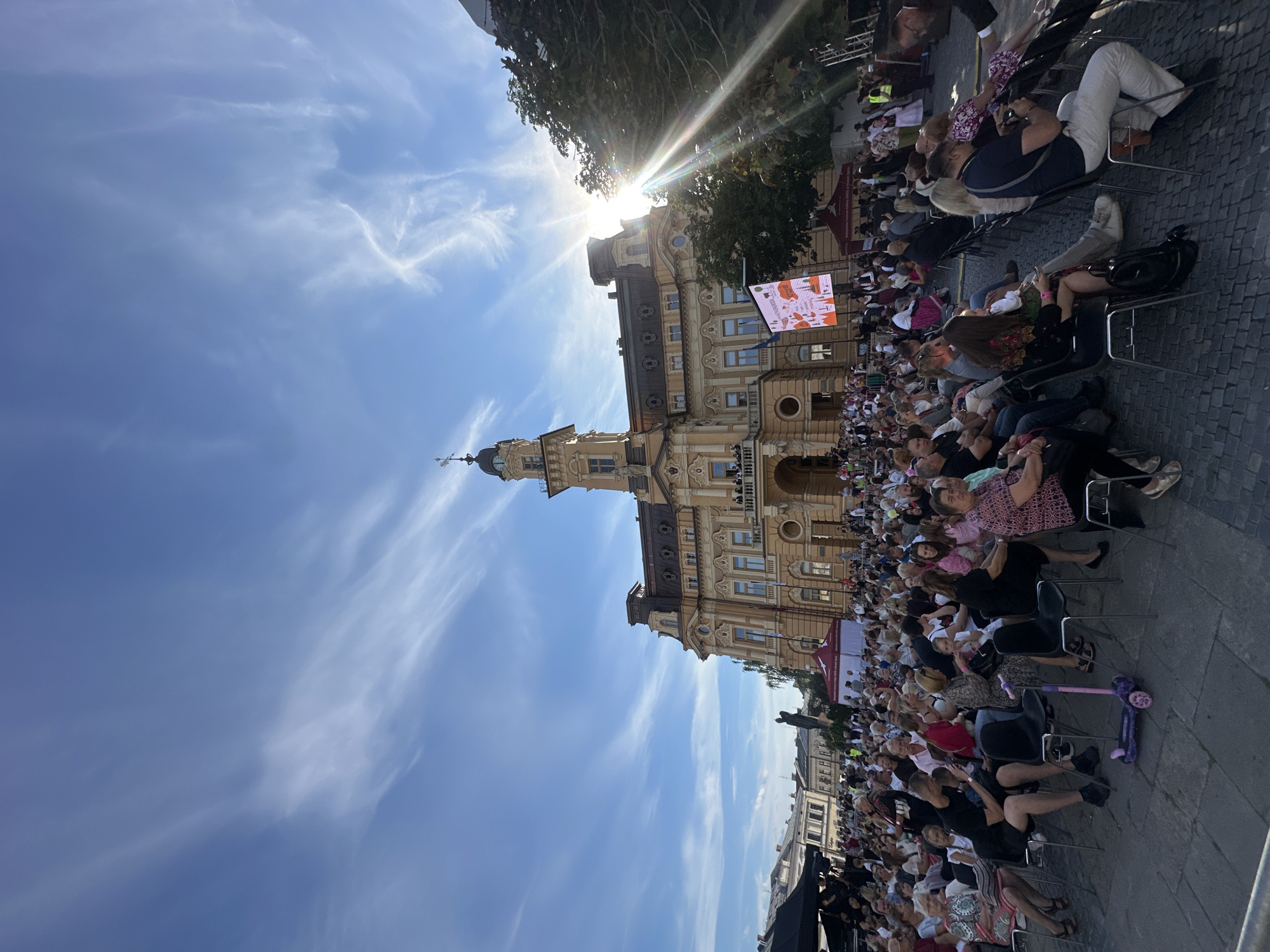 The image depicts an outdoor gathering in front of a large, ornate yellow building, identified as the City Hall in Nowy Sącz, Poland. A crowd of people, seated on chairs, are facing away from the camera, suggesting they are attending an event. The sun is shining brightly, casting lens flares and illuminating the sky, which features wispy clouds. Several tents are visible in the background, as is a large digital screen displaying an image. A child's scooter is parked on the cobblestone ground in the foreground. The architecture of the building, with its tower and decorative facade, indicates a historical and possibly important city center location. The atmosphere appears to be festive and social, with many individuals dressed in casual attire, some possibly in traditional clothing.