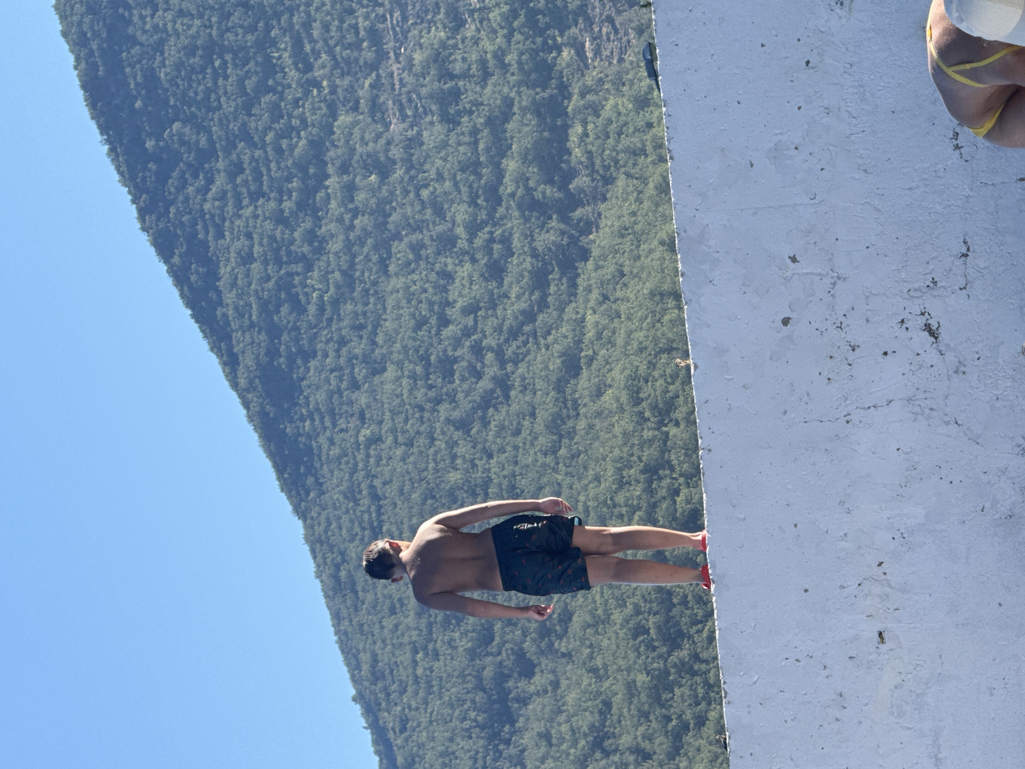 A shirtless young man with dark hair stands on a white ledge, facing away from the camera. He wears black swim trunks with a red pattern and red shoes. His body is angled towards a vast, forested mountain that rises steeply behind him, a sliver of bright blue sky visible above the mountain. To the right of the man, a white concrete structure dominates the foreground, its surface appearing rough and weathered with dark speckles. The perspective suggests the photo was taken from a high vantage point, looking down and across the scene. The lighting indicates it is daytime, likely sunny, with clear skies. A portion of another person's lower body, wearing yellow bikini bottoms, is visible at the top right of the frame, suggesting they are standing near the man on the ledge. The overall atmosphere is one of anticipation or preparation for an activity, possibly jumping or diving, given the setting and the man's stance.