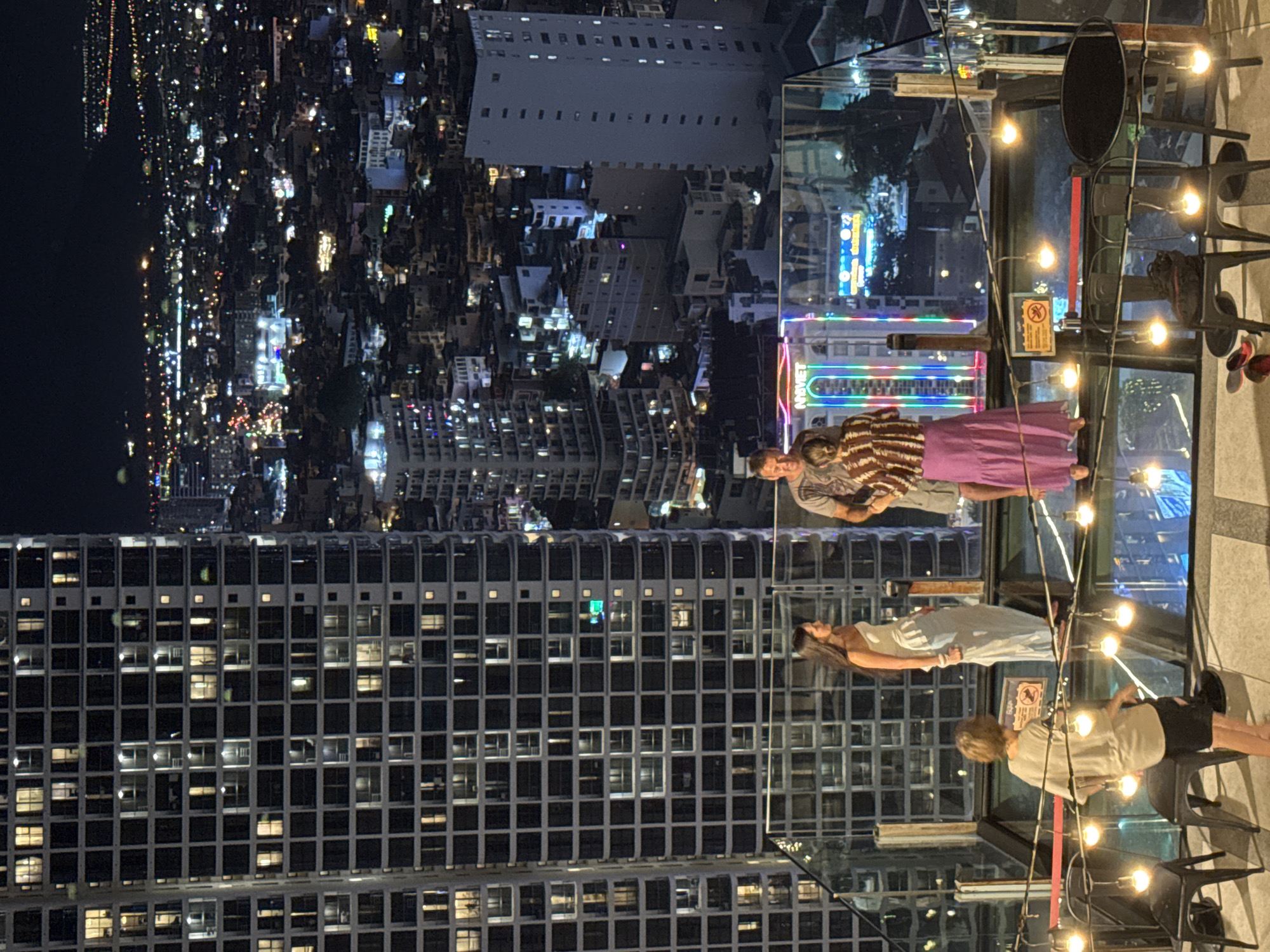 The image shows a nighttime scene on a rooftop overlooking a city in Nha Trang, Vietnam. In the foreground, a glass-railed terrace is adorned with string lights. Four people are visible: a person in a beige top and black shorts is seated, another person in a white dress is standing and looking to the side, a man in a patterned shirt and shorts is standing and facing the viewer, and a woman in a striped top and a long pink skirt is standing and looking away. Behind them, a towering skyscraper with numerous illuminated windows dominates the left side of the frame. The background reveals a densely populated cityscape illuminated by countless lights, indicative of urban development. Several other buildings of varying heights are scattered throughout the view, and a body of water is visible in the far distance. Signs with neon lights, including one with "NHAVIET" written on it, are visible in the mid-ground.