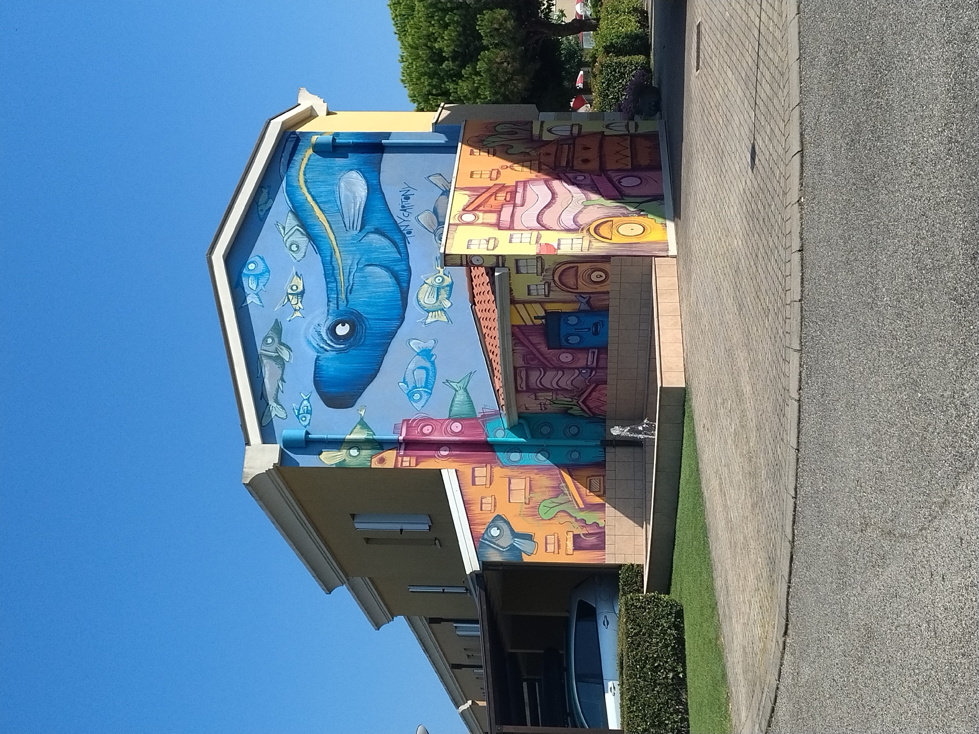 A mural depicting a blue whale and various stylized fish adorns the side of a light yellow, multi-story building. The mural extends down to a lower section of the building which features colorful, abstract depictions of buildings and geometric shapes. A small fountain is visible in front of this section. The building is situated in a paved area, with a green lawn and a hedge in the foreground. A car is parked under a carport to the left of the building. The sky is clear and blue. The mural is signed "TONY CARTONY." The scene appears to be in Caorle, Italy.
