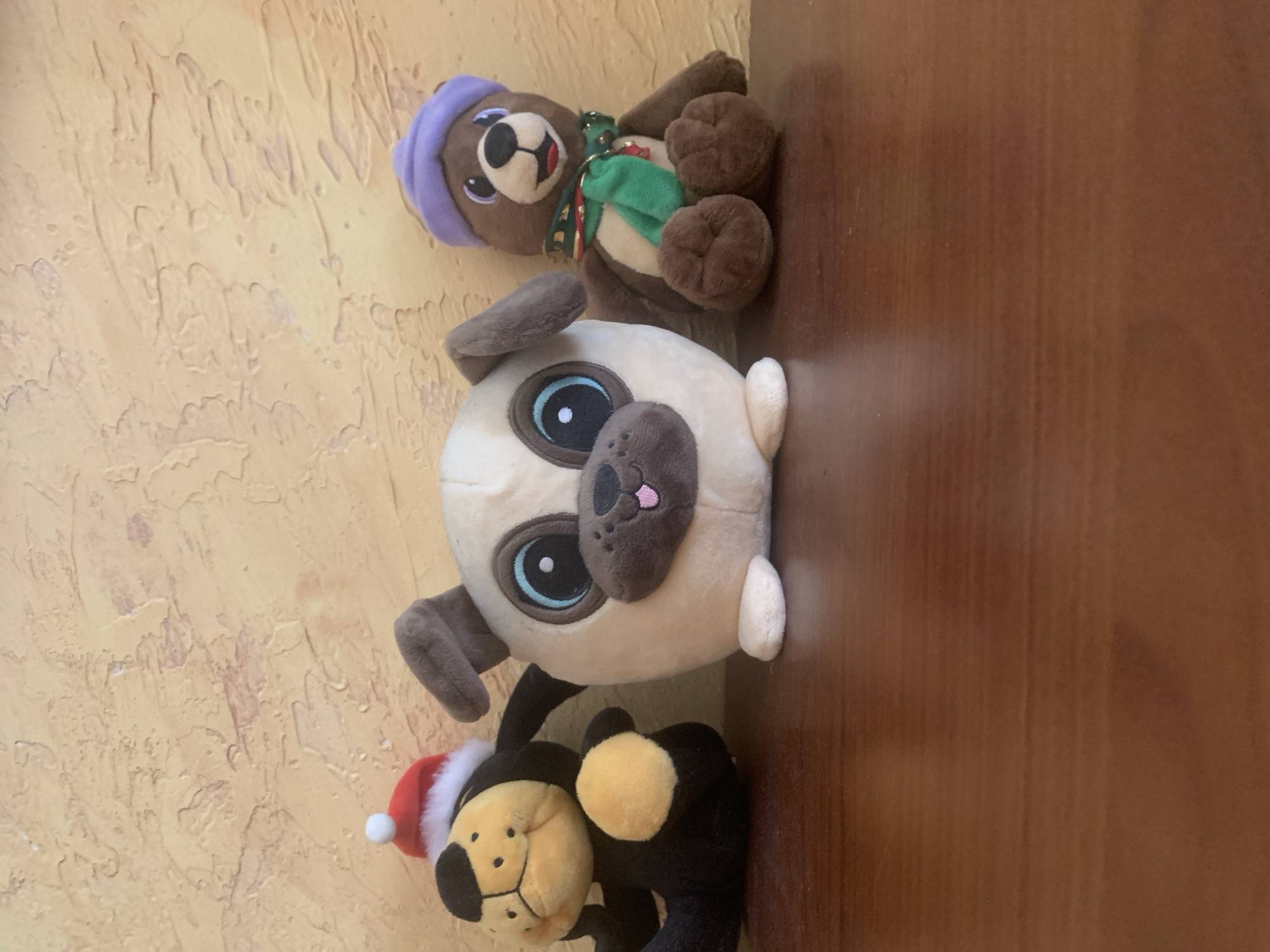 Three stuffed animals are arranged on a wooden surface against a textured, light-yellow wall. The top stuffed animal is a brown bear wearing a purple beanie and a green scarf. Below the bear is a light-tan pug with large blue eyes and dark grey ears and nose. At the bottom is a black stuffed animal with a tan face, wearing a red and white Santa hat. The animals appear to be decorative or toys, and no activity is taking place. The lighting suggests it is daytime. There is no visible text.