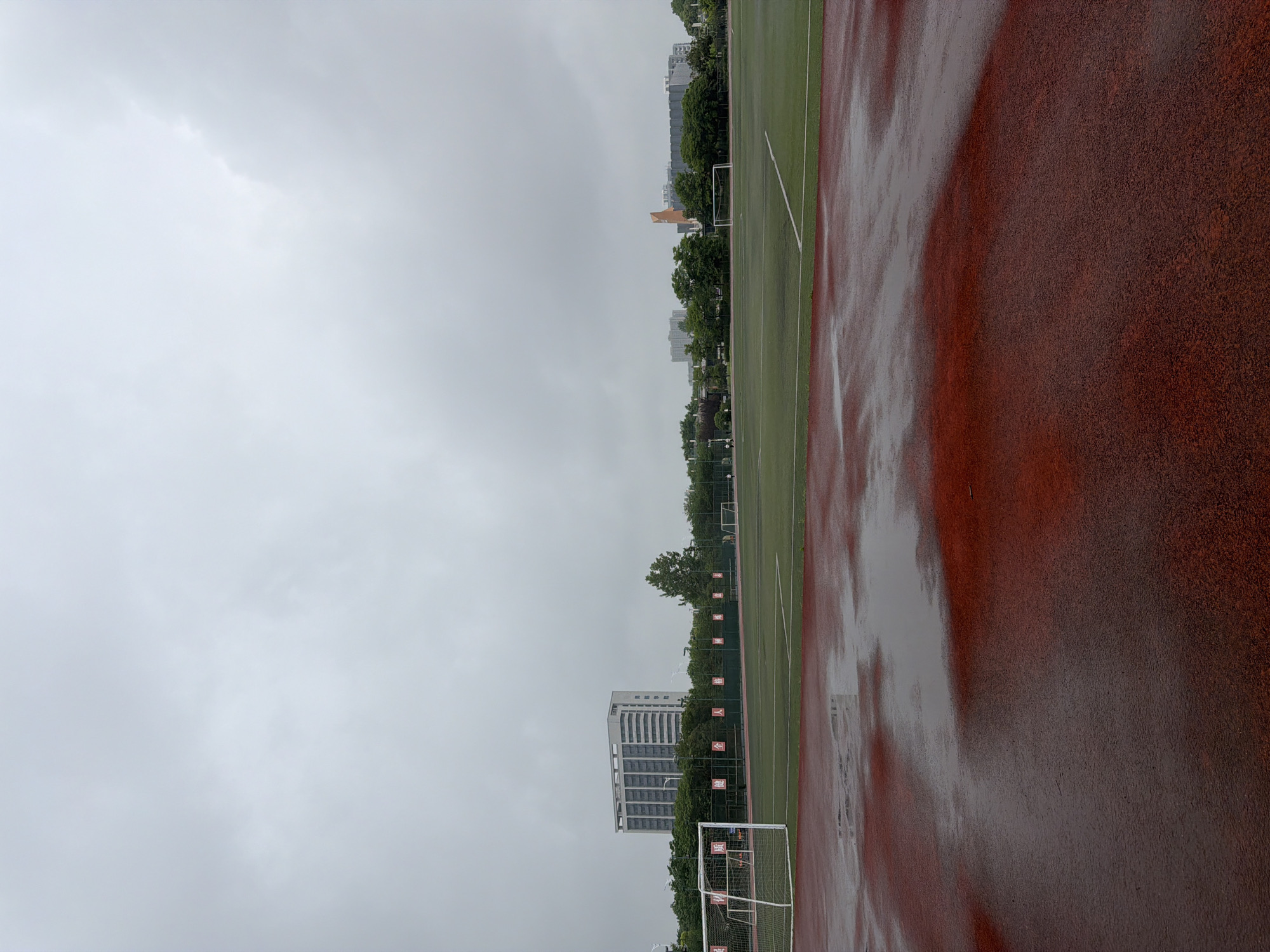 The image shows a landscape of a wet athletic field and surrounding urban environment. The sky is overcast, suggesting recent or ongoing rainfall. The athletic field is composed of a green grass area and a red running track, both visibly wet. Soccer goals are positioned on the grass field. In the background, buildings, including what appears to be an apartment complex, are visible, indicating an urban or suburban setting. Trees line the perimeter of the athletic field, and Chinese lettering flags hang. Given the prompt, the location is Jiangning District, China. There are no people visible. The scene conveys a sense of stillness due to the weather.