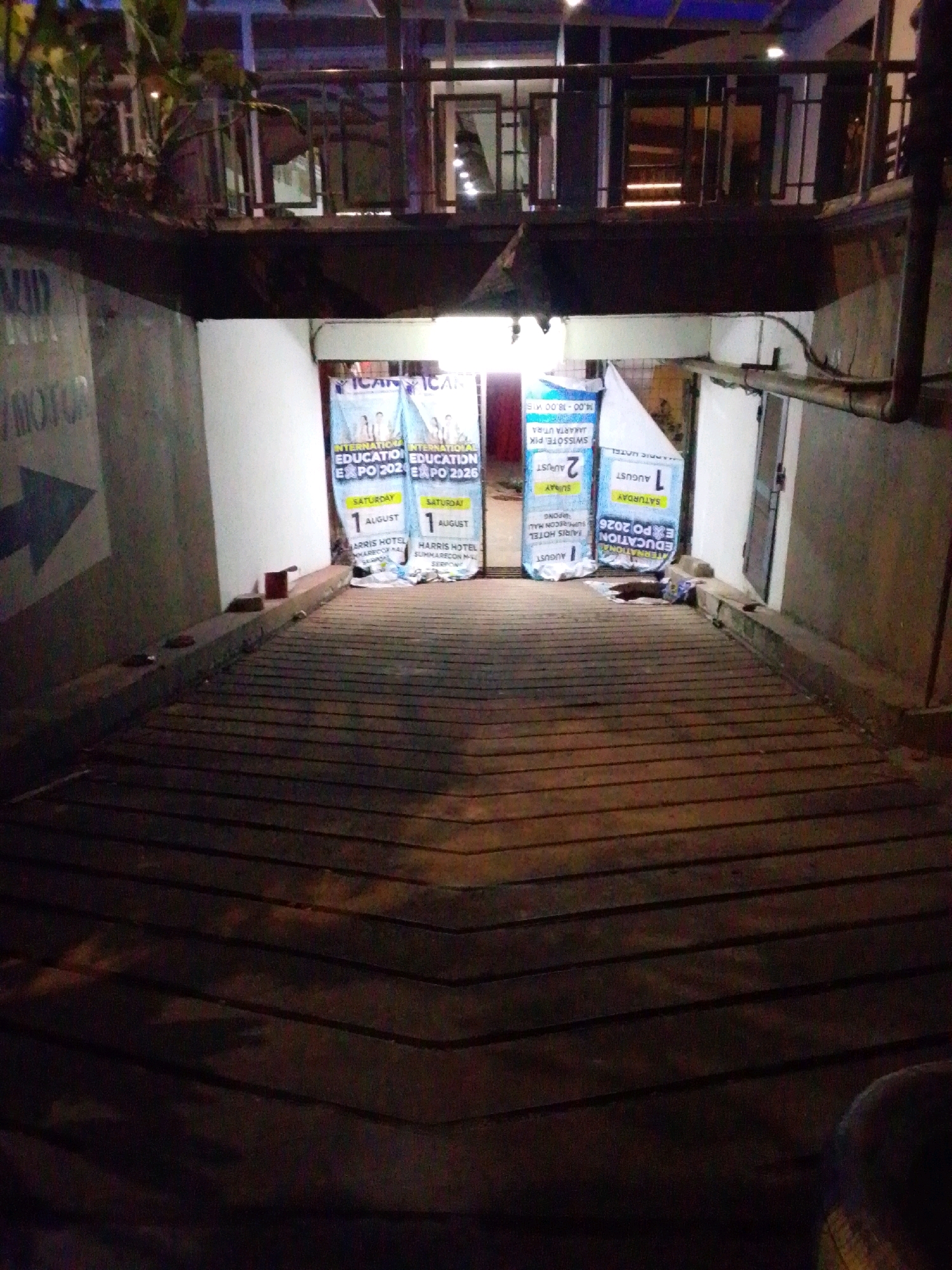 The image depicts the entrance to what appears to be a parking ramp or underground facility in North Jakarta, Indonesia. The scene is dimly lit, suggesting it is nighttime or an indoor space with limited illumination. The foreground is dominated by a sloped wooden floor, possibly a ramp, with distinct planks and shadows.
On the left side of the entrance, a concrete wall is visible, bearing a faded arrow pointing to the left and some text that is partially obscured but appears to read "MOTOR".
At the mouth of the entrance, two large blue banners are displayed vertically. The banners advertise an "INTERNATIONAL EDUCATION EXPO". The text on the banners indicates dates such as "SATURDAY 1 AUGUST" and "2 AUGUST", and mentions "HARRIS HOTEL" and "JAKARTA UTARA". The banners are slightly creased and appear to be made of fabric or vinyl.
A narrow ledge or walkway runs along the bottom of the banners and the entrance. On this ledge, several objects are scattered, including what appears to be a red brick or block, some debris, and possibly a discarded plastic bag.
The background of the image shows the interior of the structure, with lights and architectural features. There is an upper level with a railing and windows, as well as pipes and conduits running along the wall. The overall impression is of a functional, potentially commercial or event space, with the banners suggesting an upcoming event at the Harris Hotel in North Jakarta. No people are clearly discernible in the image, though there are some dark shapes at the bottom right that could be parts of individuals or their belongings.