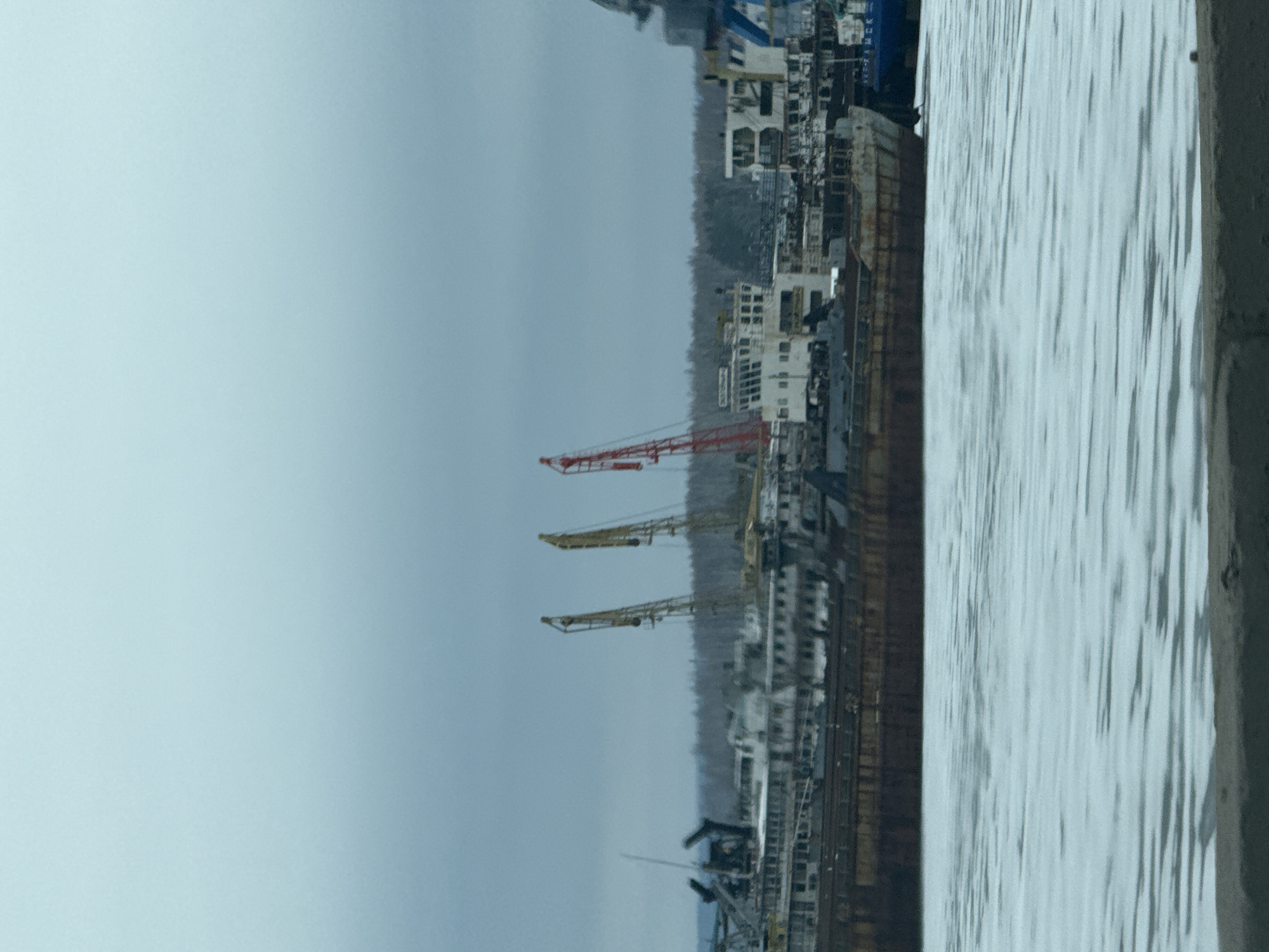The image depicts a view of the port in Chaikovsky, Russia, likely captured from an elevated vantage point. In the foreground is a body of water. Moving inland, there's a large barge docked along the shoreline, adjacent to industrial buildings and warehouses. The sky is overcast, creating a grey and muted atmosphere. Several harbor cranes are visible along the dock.
