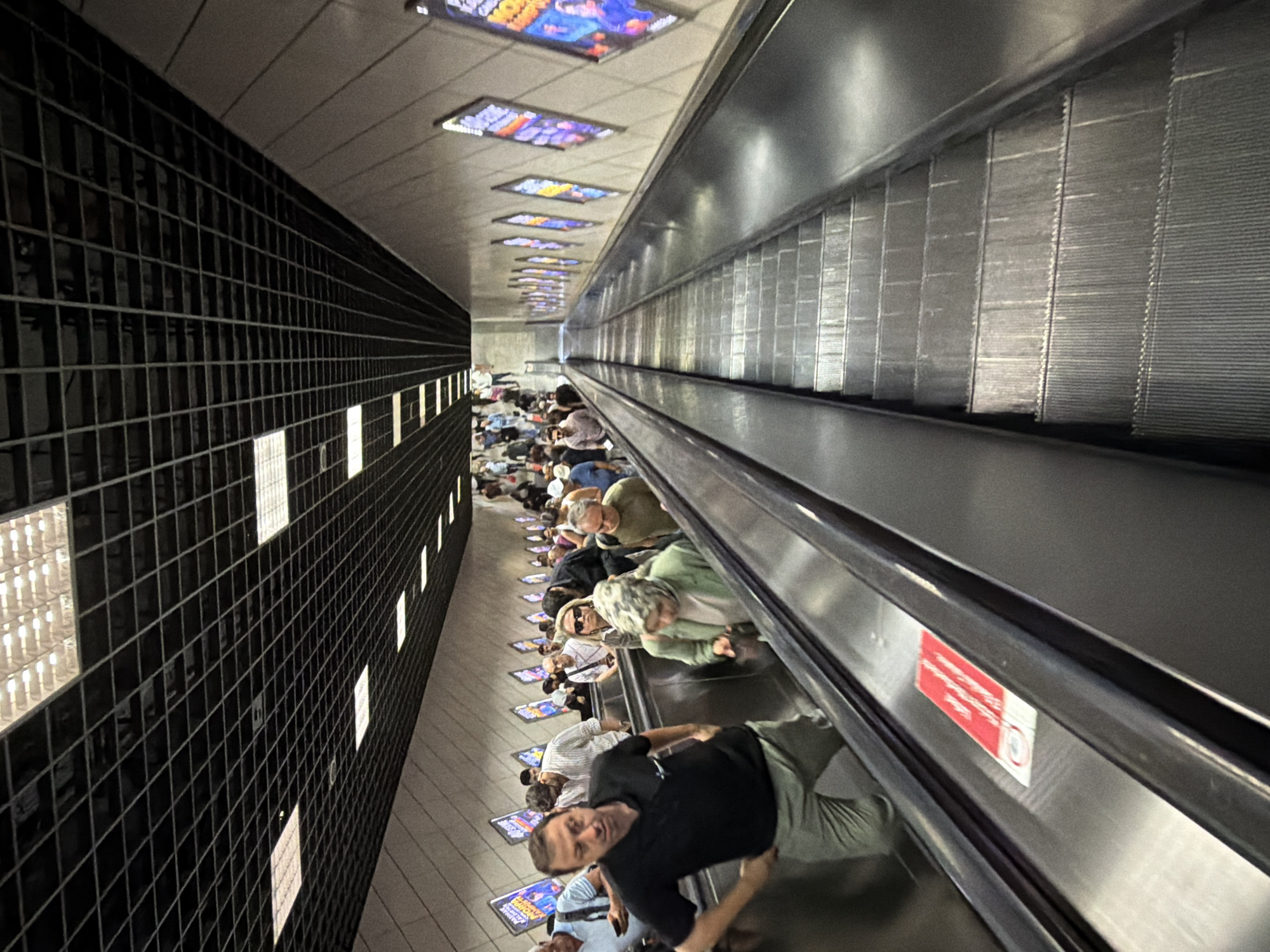 This is a street-level, eye-level, wide shot of a busy escalator in the city of Istanbul, Turkey. The escalator is going down and is filled with people of various ages, dressed in casual attire, holding bags, and appearing to be commuters or tourists. The escalator is enclosed by light-colored walls with posters and advertisements displayed periodically. Above, the ceiling is a dark, grid-like structure with recessed lighting. The scene captures the daily movement of people within the urban infrastructure of Istanbul.