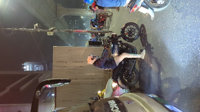 This image captures an urban street scene at night or late dusk in what appears to be Dhaka, Bangladesh. The perspective is from a vehicle, looking out onto a road with stationary traffic.
The primary subject is a person wearing a white helmet, a blue jacket, and light-colored pants, seated on a black motorcycle. The word "ROYAL" is visible on the motorcycle's fuel tank, suggesting it is a Royal Enfield. The rider's feet are on the ground, indicating they are paused. In the bottom foreground, part of a beige or white vehicle, possibly a car or van, is visible, with the word "SCANIA" partially legible on its side.
The background shows more of the city's infrastructure. An elevated road or overpass stretches across the top of the frame. Below it, several other people are present, some on bicycles or rickshaws, and others near motorcycles. Utility poles with numerous wires and a visible CCTV camera are part of the urban landscape. To the left of the main motorcyclist, there's a corrugated metal fence or wall. Further back, a brightly lit billboard displays text in Bengali script along with a portrait of a person. The scene is illuminated by artificial streetlights and vehicle lights, highlighting the dark asphalt road surface. The overall impression is a typical, bustling city street after dark.