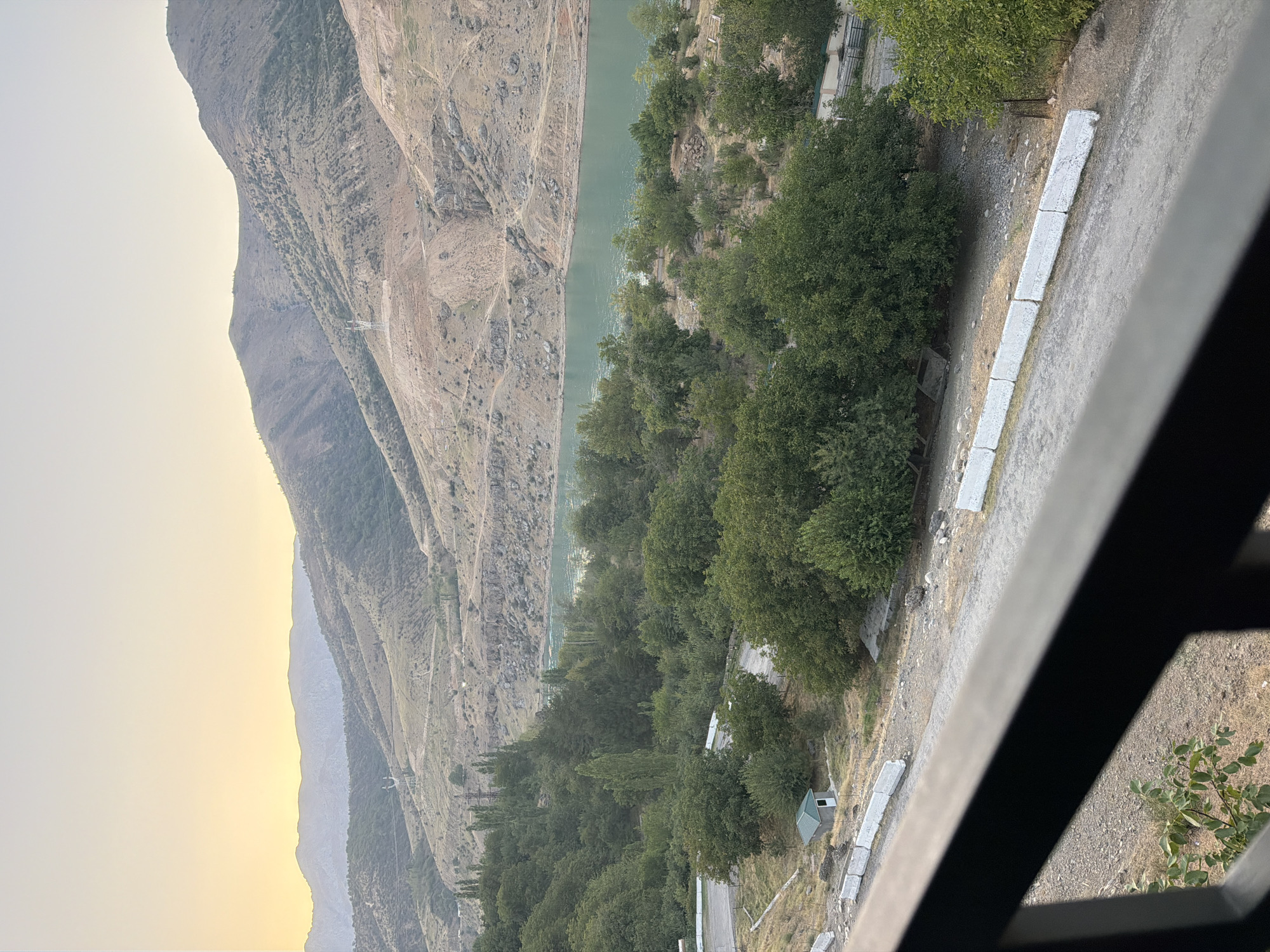 The image shows a scenic view of a mountainous landscape with a body of water. The foreground features a dark railing, suggesting the photo was taken from a balcony or similar elevated vantage point. Beyond the railing, a dirt road runs along the edge of a small, green-roofed building. Lush green trees dominate the middle ground, with a vibrant, turquoise body of water visible behind them. The background consists of rolling, barren mountains under a soft, hazy sky, possibly indicating dawn or dusk. The overall impression is one of natural beauty and tranquility. This scene is consistent with the landscape found in the Nanay region of Uzbekistan, known for its reservoirs and mountainous terrain.