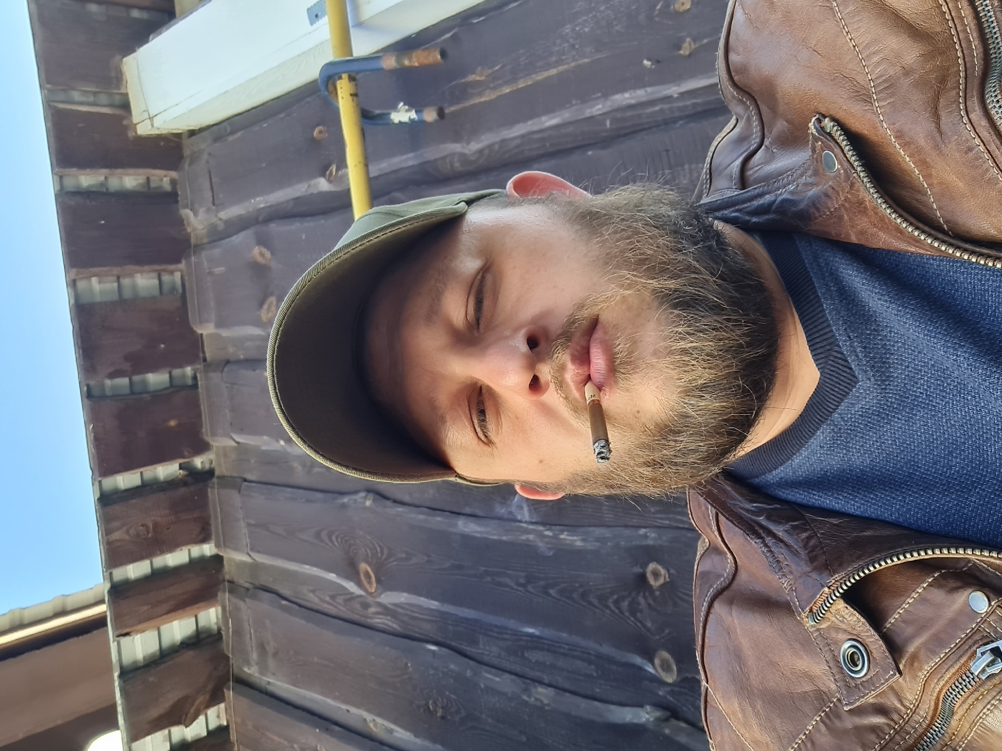 A bearded man wearing a brown leather jacket and a green baseball cap is shown smoking a cigarette. The man's eyes are partially closed. The background appears to be a wooden building with a blue sky visible in the upper left. The location appears to be Noviy, Russia.