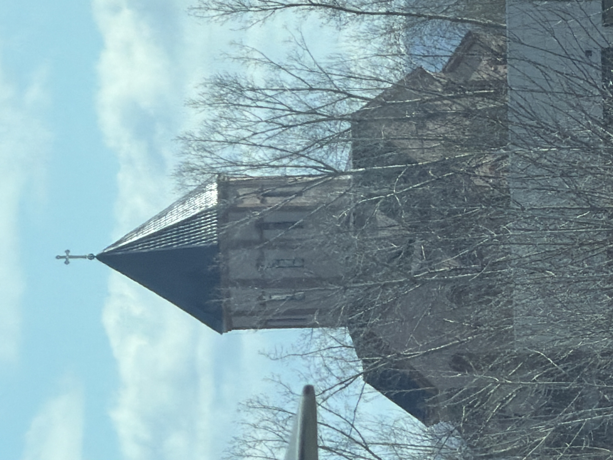 The image depicts a building in Krasnoyarsk, Russia, specifically the Cathedral of the Intercession. The cathedral is constructed of brick and has a tall, pointed steeple topped with a cross. Bare branches of trees obscure parts of the building, suggesting it is either late autumn or winter. The sky is visible in the background, partially cloudy and blue.