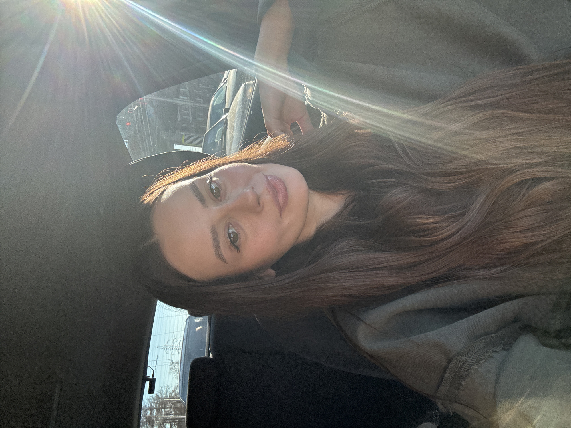 A young woman is captured in a selfie or portrait, reclining inside a car. Her long, dark brown hair cascades around her head, softly illuminated by natural light. She looks directly at the camera with a calm and slightly smiling expression, revealing light-colored eyes and defined eyebrows. She appears to be wearing a casual, muted green or grey top.
The setting is the interior of a vehicle, likely parked or moving on a street in Krasnoyarsk, Russia. Strong, bright sunlight streams in from an overhead window, creating prominent lens flares and visible sun rays across the top left of the image, indicating daytime. Through the car windows, a blurred background of an urban or suburban environment is visible, including other vehicles, buildings, and utility structures in the distance. The car's interior consists of dark-colored fabric. No discernible text is visible in the image.