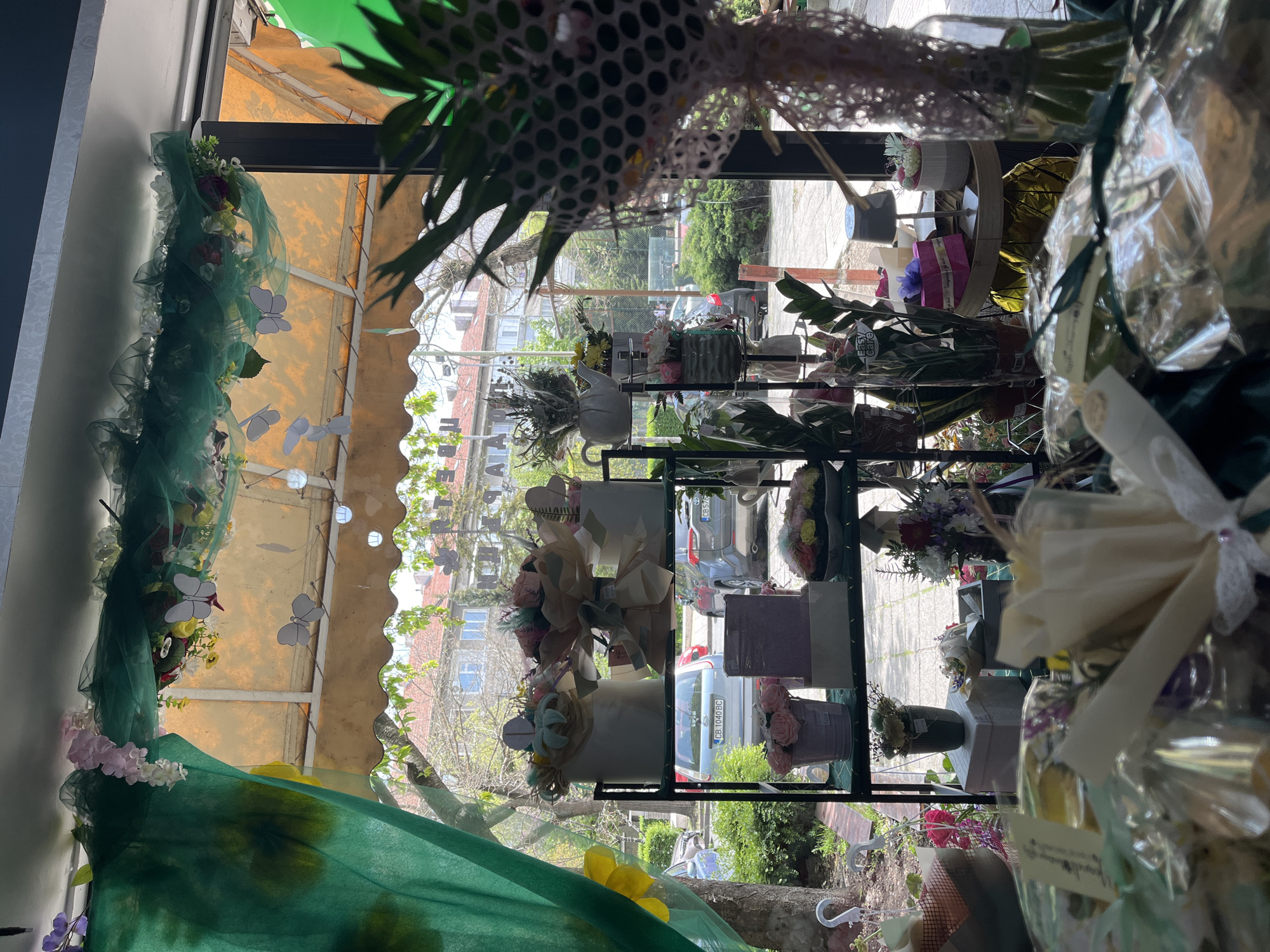 The image depicts the interior of a flower shop, seemingly located in Burgas, Bulgaria, as inferred from the reflection in the glass showing a license plate with "OB 1040 BC" which follows the Bulgarian vehicle registration code. A metal shelf displays a variety of floral arrangements in containers and wrapped bouquets. The window display features green tulle fabric with decorative butterfly accents. Outside, a street scene is visible with cars parked along the curb, trees lining the sidewalk, and buildings in the background.