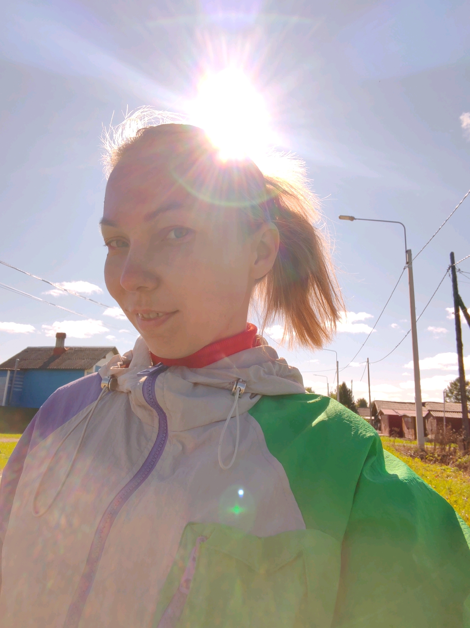 A young woman with blonde hair tied in a ponytail takes a selfie outdoors on a sunny day. She is wearing a multi-colored windbreaker, predominantly white with green and purple accents, and a red collar is visible underneath. The sun is high in the sky, creating lens flare and washing out some details. In the background, a blue house with a brick chimney is visible to the left, and a street light and power lines stretch across the frame. The scene appears to be in a rural or semi-rural area, possibly a village in Чухлэм, Russia, given the presence of simple houses and utility poles. The overall impression is a casual, outdoor snapshot, with the bright sunlight being a dominant feature of the composition.