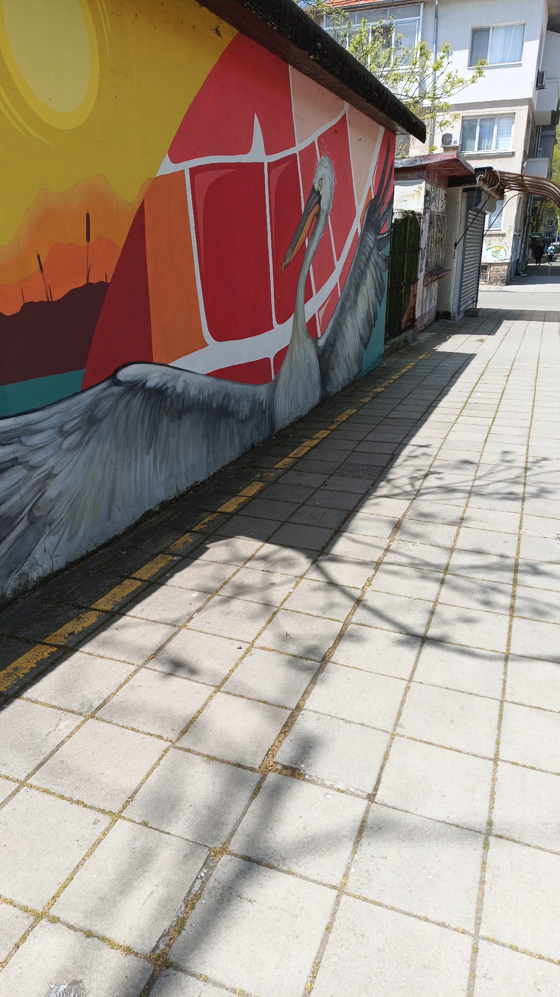 The image shows a sidewalk in Burgas, Bulgaria, with a mural painted on the side of a building. The mural depicts a pelican against a background of colorful geometric shapes and a sunset scene. The sidewalk is paved with square concrete tiles, and there are shadows cast by trees. The image includes some building structures.