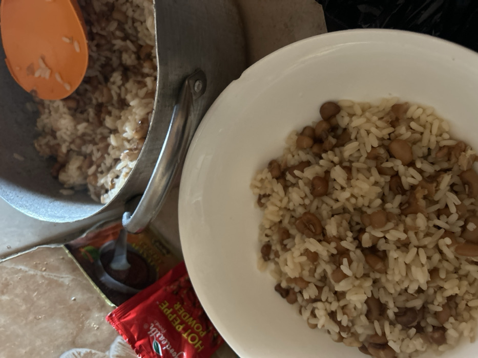 The scene, located in Egbe, Nigeria, depicts a prepared meal. In the foreground, a white bowl contains a serving of cooked white rice intermingled with brown beans. In the background, a metal pot holds a larger quantity of the identical rice and bean mixture, with an orange plastic spoon partially visible within the food. To the left of the pot, a red packet labeled "goodearth foods HOT PEPPE POWDER" rests on a light-colored, possibly tiled surface. A partially obscured yellow-brown packet is situated behind the red one. A dark, indistinct object is visible in the bottom right quadrant. No individuals are present in the image.