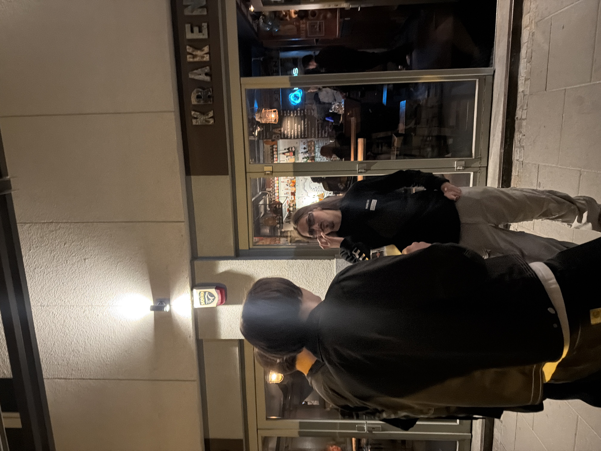 The image captures a nighttime street scene in front of a bar named "Kraken". Two people are visible in the foreground, with one person looking towards the camera. The other person is looking down and to the side. In the background, the bar is open and populated with patrons. A light shines on the wall to the left, near a sign. The scene is urban, likely taking place in the city.
