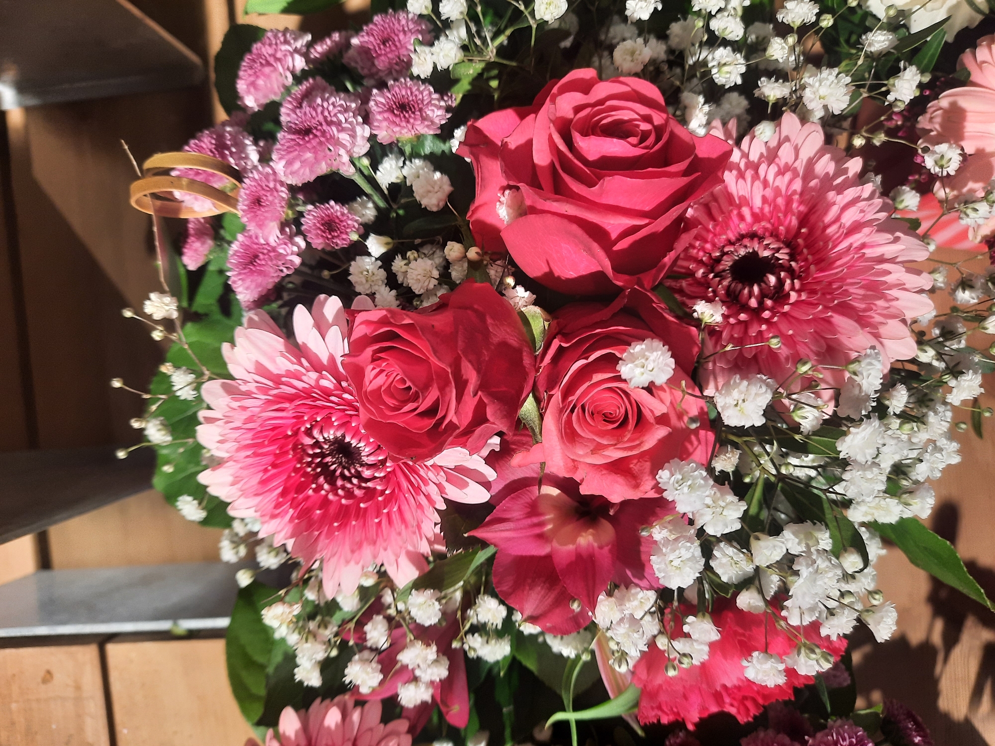 A close-up, eye-level shot captures a vibrant bouquet of pink and red flowers. Dominating the foreground are large pink Gerbera daisies with dark centers, interspersed with deep pink roses showing their layered petals. Smaller, clustered pink flowers, possibly chrysanthemums, add texture to the arrangement. Delicate sprigs of white baby's breath are scattered throughout, providing a airy contrast. A hint of red carnation can be seen on the left. Two gold wedding rings are subtly placed near the top center of the bouquet, tied together by a thin brown string, suggesting a wedding or anniversary occasion. The background is blurred, showing wooden elements and a metallic structure, hinting at an indoor setting. The lighting suggests a bright, possibly sunny day, casting soft shadows. The image does not depict any specific location in Rosenheim, Germany, but rather a generic floral arrangement.