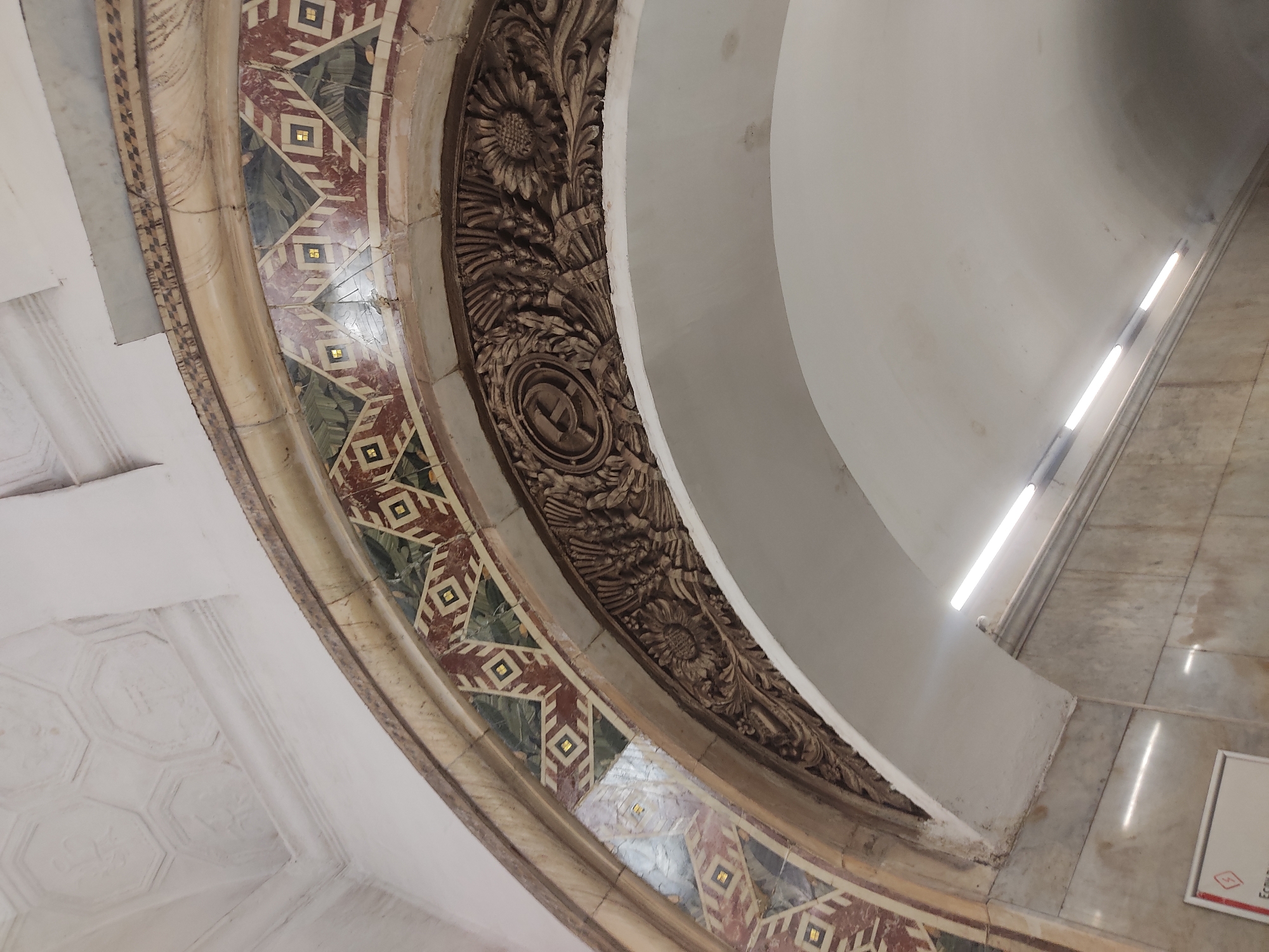 The image shows a section of the ceiling architecture of the Kievskaya Metro Station in Moscow, Russia. The architectural details include an elaborately carved frieze with floral motifs and the symbol of the Soviet Union (hammer and sickle) incorporated into the design. The border is decorated with geometric patterns and contrasting colored stones. To the right, there is a smooth, white curved portion of the ceiling, illuminated by built-in light fixtures. A marble floor with some reflections is visible, and a sign is partially visible.