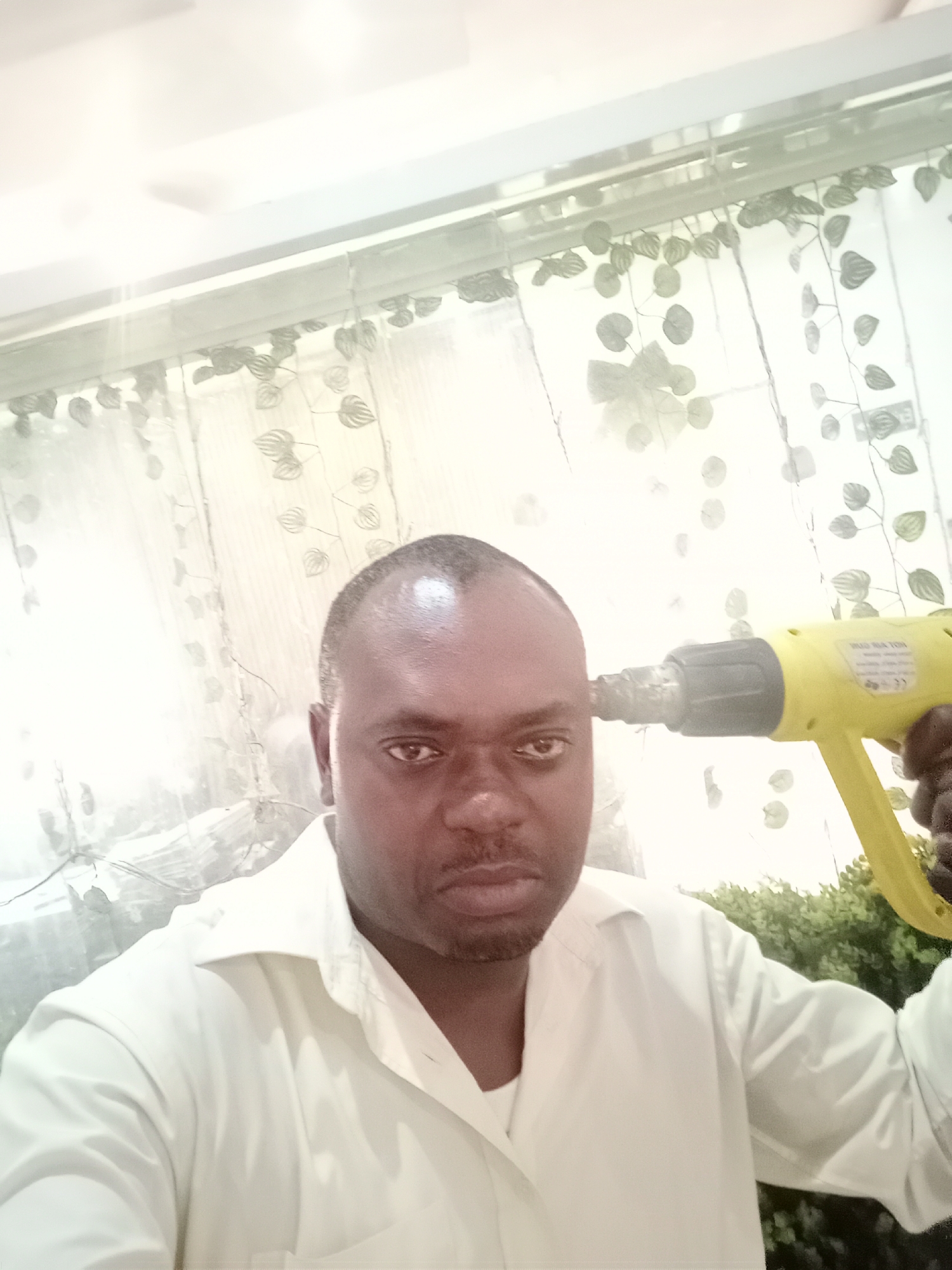 An adult Black man is pictured indoors, holding a yellow heat gun to his head, presenting the illusion he's shooting himself. He is wearing a white button-down shirt. The background includes a window with sheer curtains with a leaf pattern. Behind the window, greenery is visible. The location may be in Gwarinpa, Nigeria.