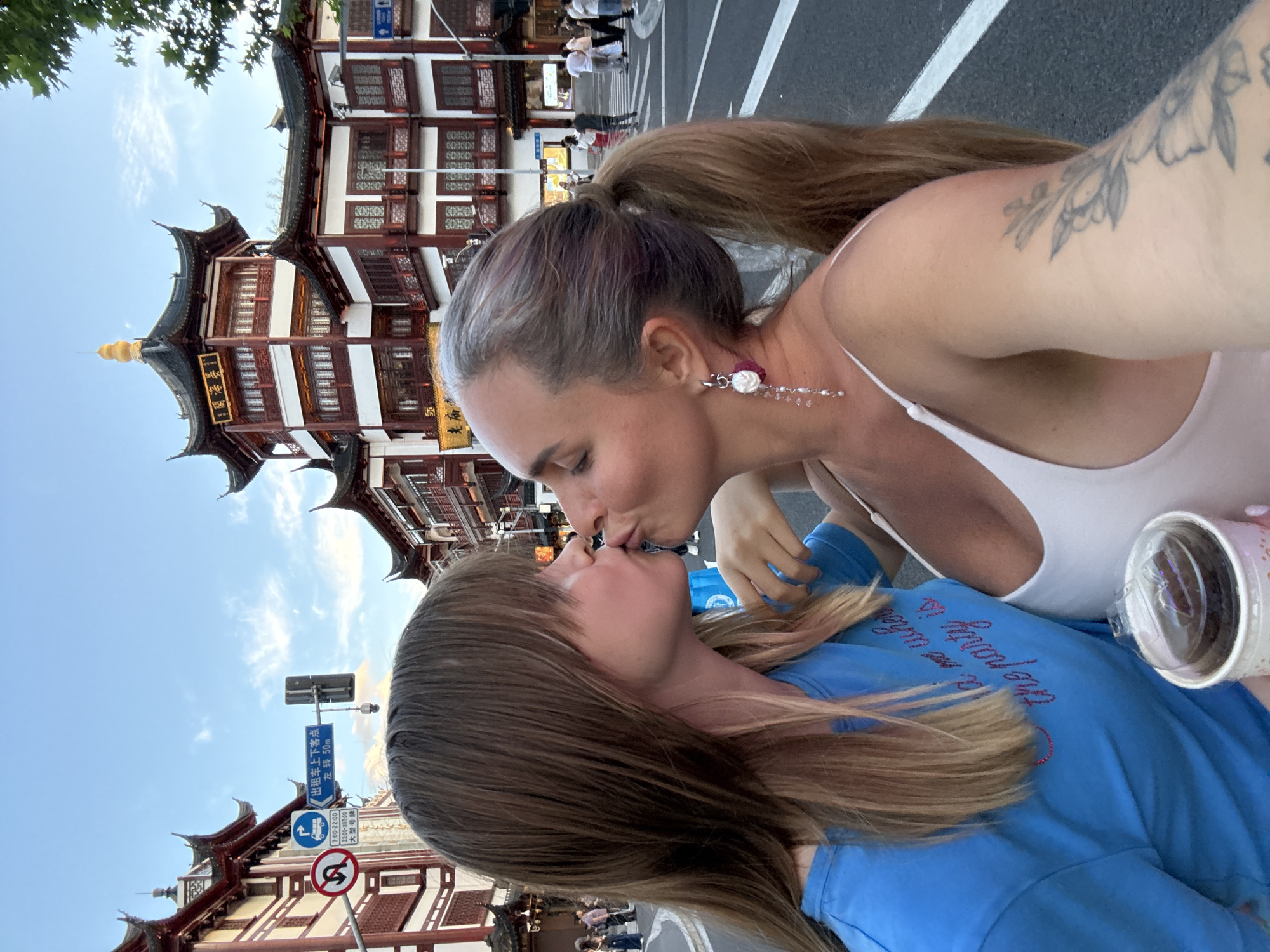 In Shanghai, China, two women are captured in a close-up selfie as they share a kiss. They are standing on a street with traditional Chinese architecture in the background, characterized by multi-tiered roofs with ornate details and vibrant red and white facades. The sky is a pale blue with scattered clouds.
The woman on the left has long, light brown hair with bangs and is wearing a bright blue t-shirt with some text that is partially visible. The woman on the right has her hair pulled back in a ponytail and is wearing a white tank top. She has a long, dangly earring with beads and pearls. A tattoo of floral elements is visible on her arm.
In the foreground, there is a white cup with a clear lid containing a dark-colored dessert, possibly a cupcake or mousse. The street scene indicates a bustling urban environment with a pedestrian crossing and traffic lights visible. Several other people are present in the background, walking along the street. A street sign with Chinese characters and directional arrows is also visible.