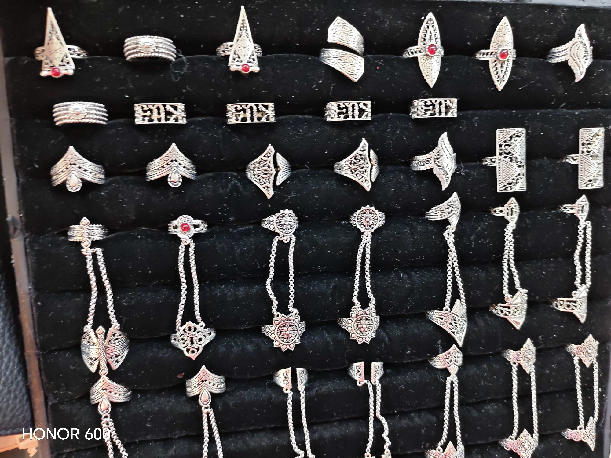 The image displays a black velvet-lined display case filled with various silver-colored rings and hand jewelry. The jewelry features intricate, ornate designs, some incorporating small red stones. The items are arranged in rows. The bottom left corner of the image contains the text "HONOR 600". The scene appears to be a retail display of accessories. There is no information within the image to suggest a location in Ussurijsk, Russia.