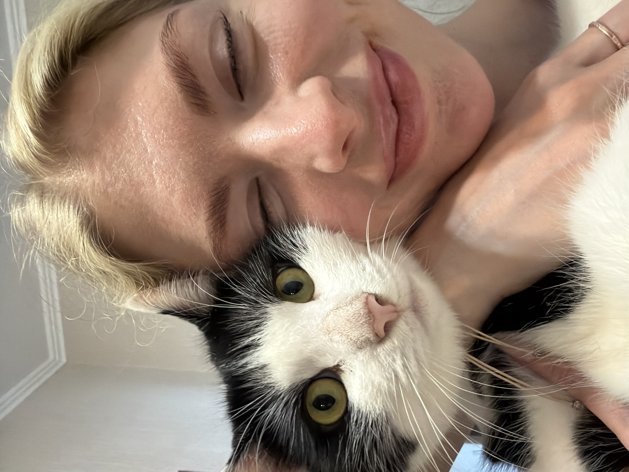 The image is a close-up selfie featuring a young blonde woman and a black and white cat. The woman is on the right side of the frame, with her eyes closed and a slight smile. She is wearing a white top. The cat is on the left side of the frame, its head resting on the woman's arm and shoulder. The cat has green eyes, a pink nose, and black and white fur. The background is blurred and appears to be an indoor setting, possibly a room with light-colored walls and molding. The image is taken from a slightly high angle. No specific location in Khimki, Russia is identifiable from the visual information.