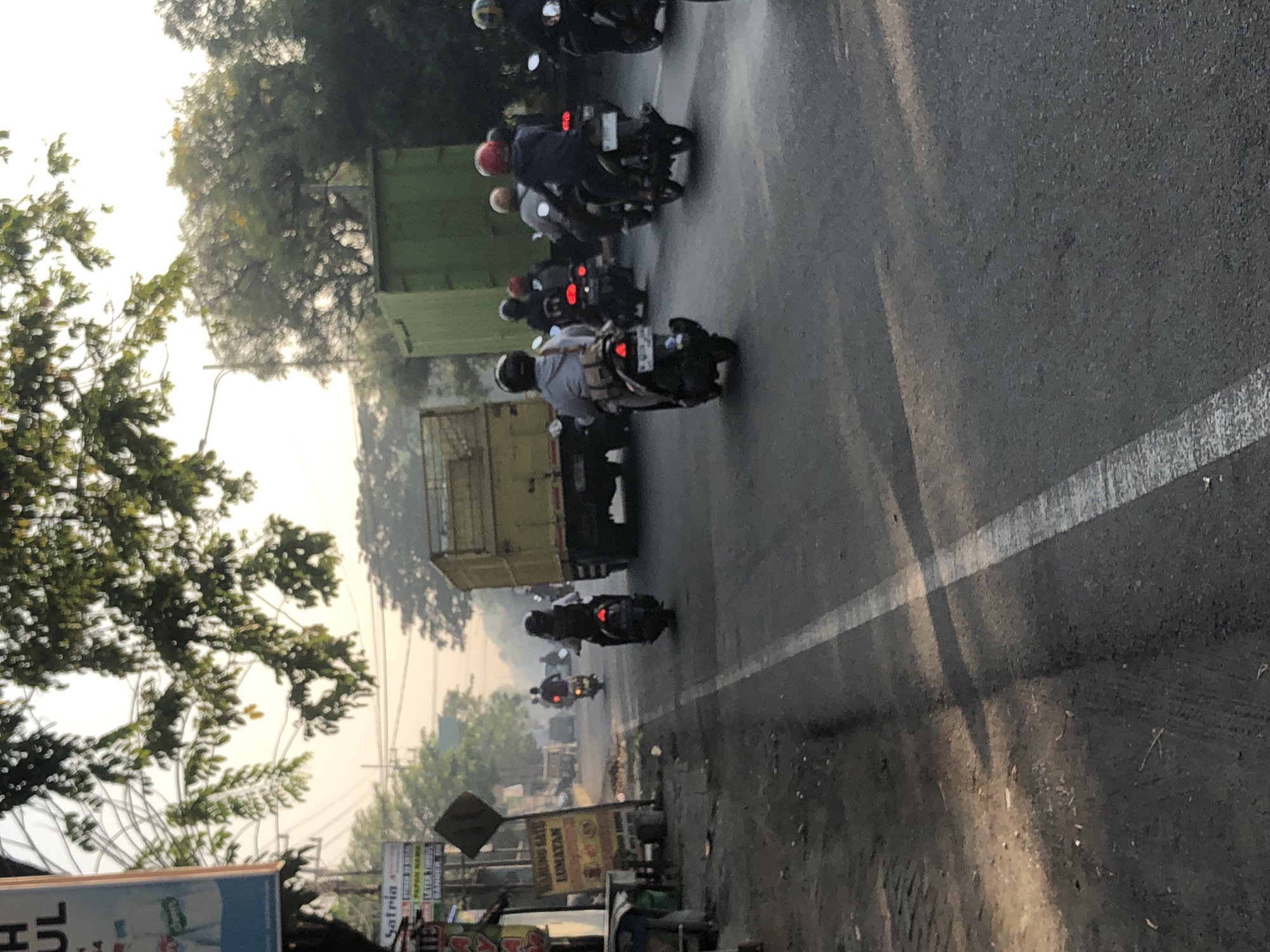 This image captures a street scene in Kludan, Indonesia, characterized by a line of motorcycles and a truck on a paved road. The sky is overcast, suggesting it might be early morning or late afternoon. Lush green trees line the sides of the road, with some branches extending over the street. The road has a white dividing line and appears to be a busy thoroughfare, with several motorcycles in motion.
In the foreground, a motorcycle with a rider wearing a grey hoodie and a backpack is visible. Further down the road, more motorcycles are seen, some carrying passengers. A large yellow truck is positioned on the right side of the image, contributing to the sense of traffic flow. Signs are visible on the left side of the road, indicating commercial activity. The overall impression is that of a typical day on a road in a bustling Indonesian town or city.