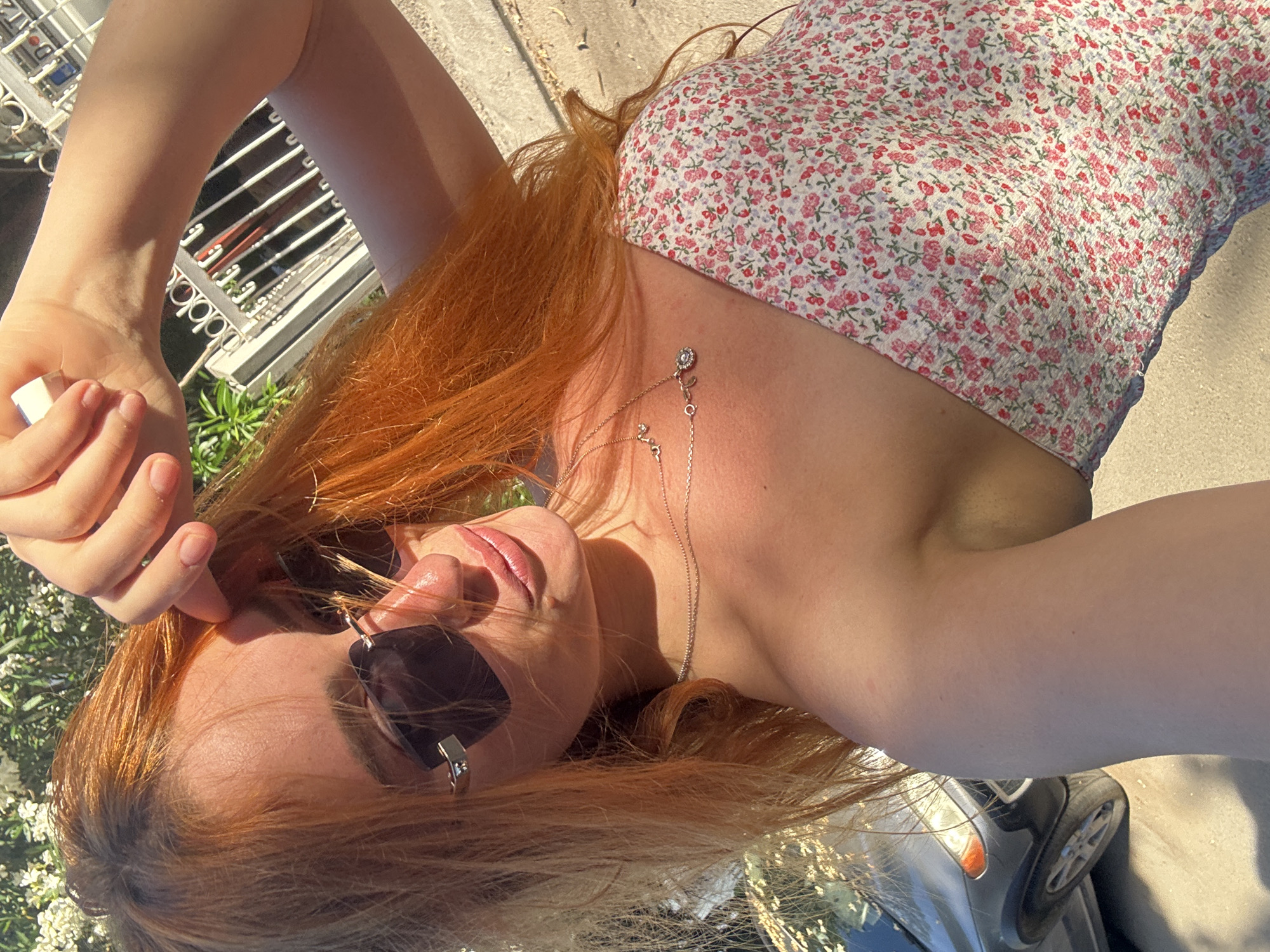 A female with long, reddish-orange hair is depicted in a selfie, likely taken outdoors in Budva, Montenegro, under bright sunlight. She is wearing dark sunglasses and a light-colored strapless or off-the-shoulder dress with a small floral pattern of red and pink blossoms. Two delicate silver-toned necklaces are visible on her chest, one appearing to have multiple small charms and another with a single, possibly circular, pendant. Her right arm is raised, with her hand positioned near her forehead, possibly adjusting her hair or shading her eyes. The background features green foliage, some with white flowers, and a white metal fence. A glimpse of a dark-colored car with a visible wheel and headlight is seen in the lower left, on what appears to be a paved surface or road. The overall lighting suggests a clear, sunny day.