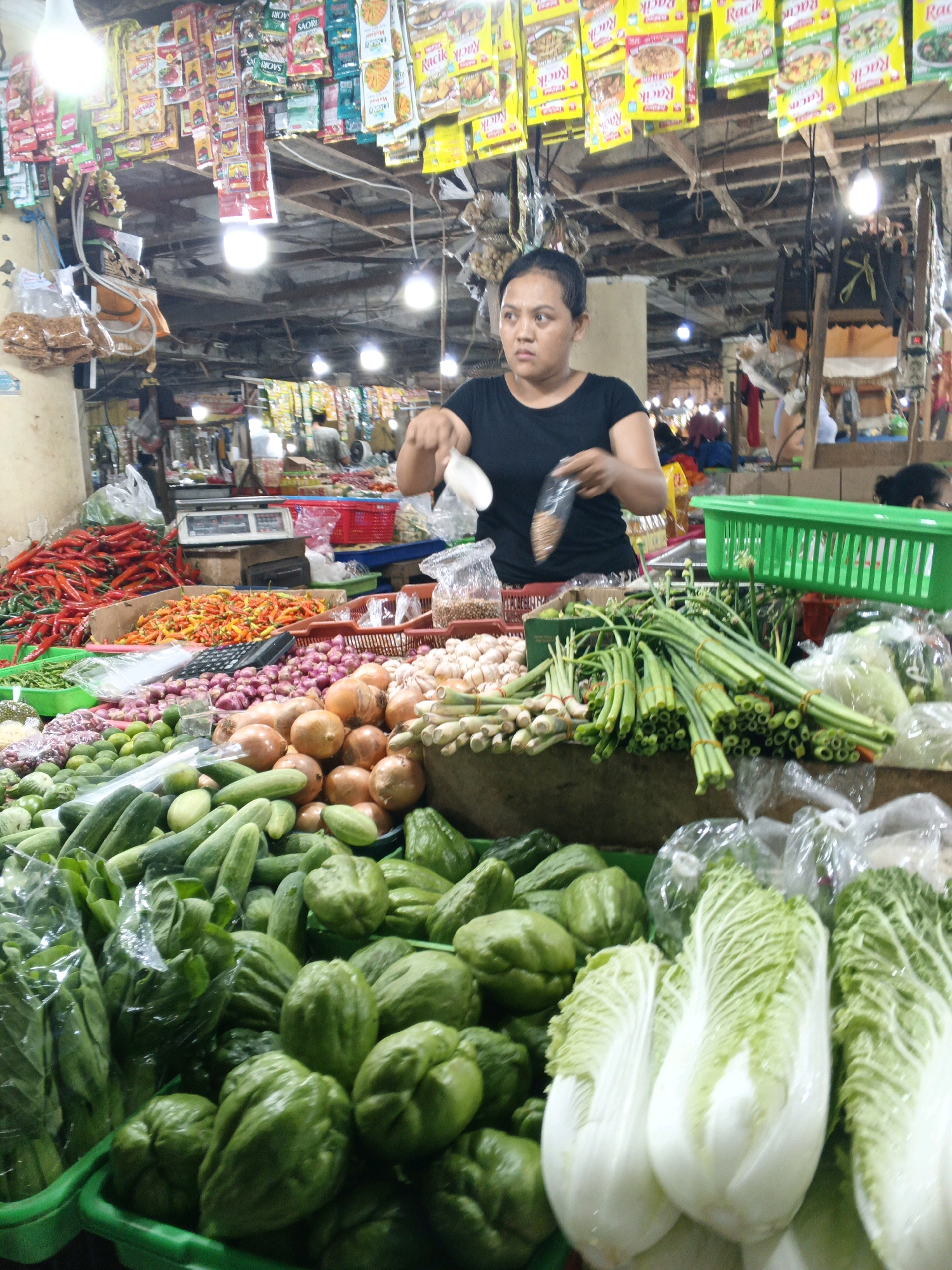 A young woman, wearing a black t-shirt, stands behind a stall in a market, holding a clear plastic bag filled with dried goods. The stall is laden with a variety of fresh produce, including piles of red and yellow chilies, onions, garlic, cucumbers, and leafy green vegetables like bok choy and Napa cabbage. Behind the woman, shelves are stocked with numerous brightly colored food product packages, many of which are identifiable as seasoning packets. The market appears to be covered, with overhead lighting illuminating the scene. The overall atmosphere suggests a typical Indonesian market environment, likely in Denpasar, Indonesia, given the prevalence of such bustling local markets in the city.