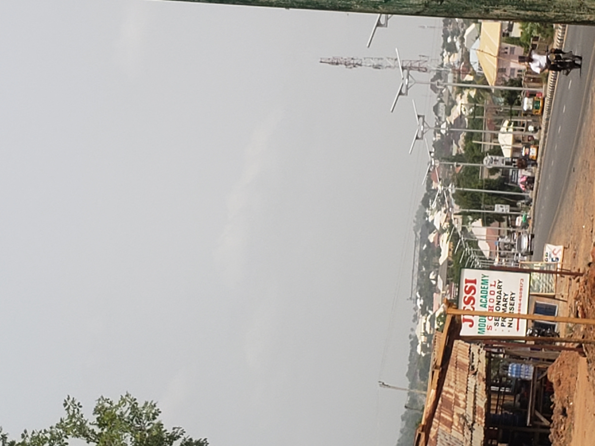The image depicts a street scene in Adankolo, Nigeria. The road runs horizontally across the frame, lined with solar-powered street lights. A motorcycle with two riders is traveling along the road. The background consists of buildings, greenery, and what appears to be a construction crane.
In the foreground, a sign for "Jessi Model Academy School" is visible beside a partially obscured building structure with a corrugated metal roof. Water bottles are stacked near the building. The sky is overcast.