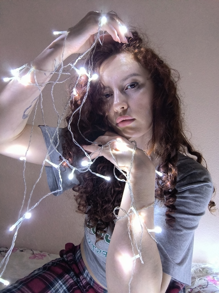 The image portrays a female subject in an indoor setting. She has long, curly, reddish-brown hair and looks directly at the camera with a neutral expression. A string of illuminated white LED lights is draped across her upper body, arms, and hair. Her left arm is raised, with the lights wrapped around it and through her hair; a dark, stylized tattoo is visible on her left upper arm. Her right hand rests near her chin, also entwined with the lights. She is dressed in a grey short-sleeved crop top and red plaid pajama bottoms. A floral-patterned textile, consistent with a bedsheet, is visible in the lower foreground. While the requested location is Saint Petersburg, Russia, the image contains no visual identifiers to confirm this geographical context or a specific location within the city.