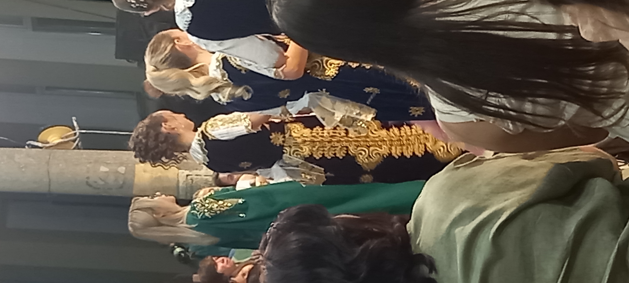 The image depicts a group of people, primarily women, dressed in traditional attire. The focus is on several individuals wearing elaborate outfits with intricate gold embroidery. One woman on the left wears a flowing green garment with gold accents on the shoulders. In the center, a woman is seen from behind, wearing a dark vest over a white, gold-trimmed blouse and a skirt or dress with extensive gold ornamentation and sheer panels. Another woman to her right also wears a similar dark vest and gold-trimmed blouse. A person with blonde hair pulled back in a bun is visible in the center. The background appears to be an outdoor setting, possibly a courtyard or street, with a concrete pillar on the left and what seems to be a building structure in the distance. The lighting suggests it is daytime, with some shadows present. The overall impression is of a cultural event or performance. The context of Lyuben, Bulgaria, is not visually discernible from the image itself.