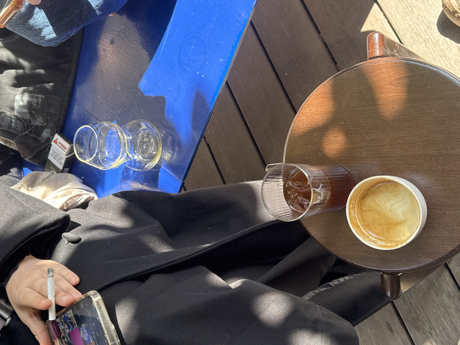 The image depicts a close-up, high-angle view of an outdoor cafe or restaurant setting in Tel-Aviv, Israel, identifiable by the Hebrew health warning on a cigarette pack.
In the foreground, a round, dark brown wooden table supports a clear, ribbed glass containing a dark beverage with ice cubes (likely iced coffee) and a white ceramic cup holding light brown coffee residue with remnants of latte art. Strong, direct sunlight casts distinct shadows across this table and the wooden plank flooring.
To the left, a person is partially visible, wearing a dark coat. Their left hand holds a lit cigarette with an ash tip and a smartphone in a clear case. The phone screen displays multiple small image thumbnails and the partially visible word "Favorites."
In the middle ground, a vibrant blue table, showing signs of wear with a chipped edge on its right side, holds an empty, clear, hourglass-shaped glass. A Marlboro Red cigarette pack with a Hebrew health warning is visible behind the glass, next to a dark, textured bag or garment. A portion of a second person's leg, clad in blue jeans, is visible on the far right of the blue table, suggesting another individual is present. The ground consists of dark wooden planks.