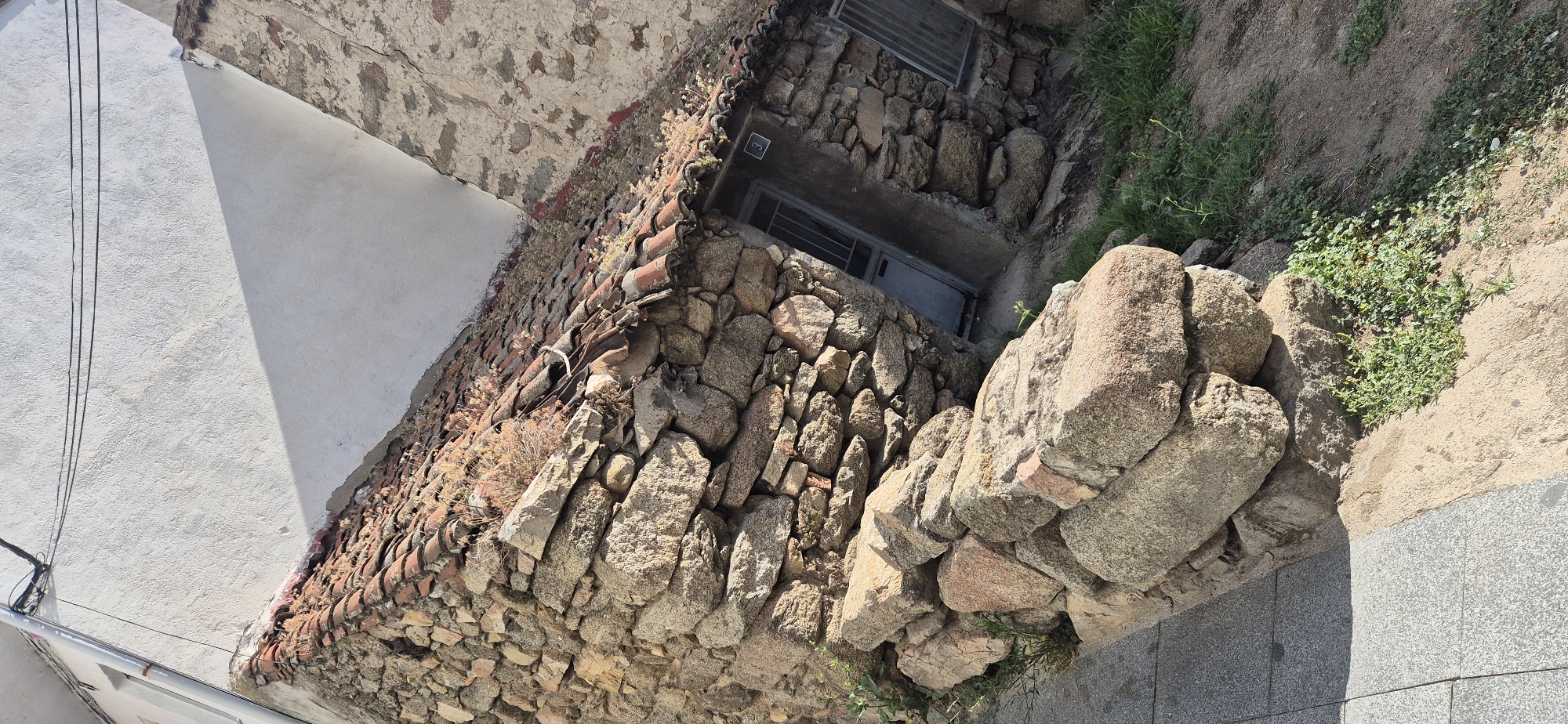 A narrow, paved walkway runs along the bottom left of the image, bordered by a rough, stacked stone wall. This wall is composed of large, irregular stones of varying shades of brown and gray, some appearing weathered and rounded. Behind the stone wall, the roof of a small, rustic structure is visible, covered in weathered, reddish-brown terracotta tiles and overgrown with dry vegetation. The structure has a small, dark doorway with a metal grate and a small metal grate window to its right. The building is constructed of similar rough stones as the wall. To the upper right, a white, textured wall of another building casts a sharp triangular shadow across the scene. Electrical wires are visible strung across the top of the white wall. The ground to the right of the structure is a sandy, dirt surface with patches of green grass and weeds. The scene appears to be an outdoor, possibly rural or older urban, setting under bright daylight conditions, evidenced by the strong shadows. There are no people or animals visible in the image. The number "3" is faintly visible above the doorway.