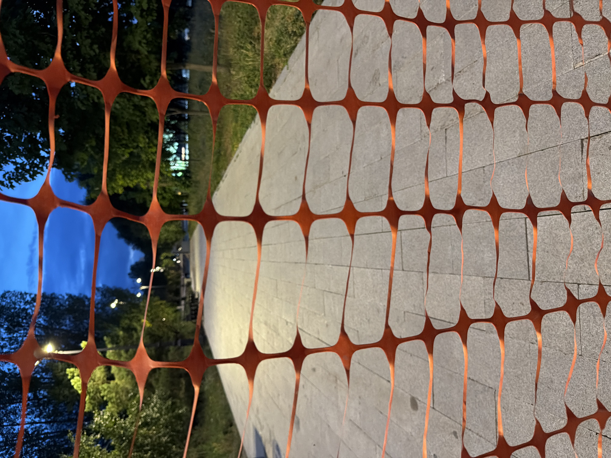 The image shows an orange plastic fence in the foreground, obscuring a paved pathway in an urban park. The pathway is made of light-colored rectangular pavers and is lined with grass and trees. In the background, streetlights are visible, indicating that it is dusk or night. The sky is a deep blue. The scene appears to be located in Astana, Kazakhstan, as suggested by the style of the pathway and streetlights, which are common in urban parks in the region. There are no people or animals visible in the image.