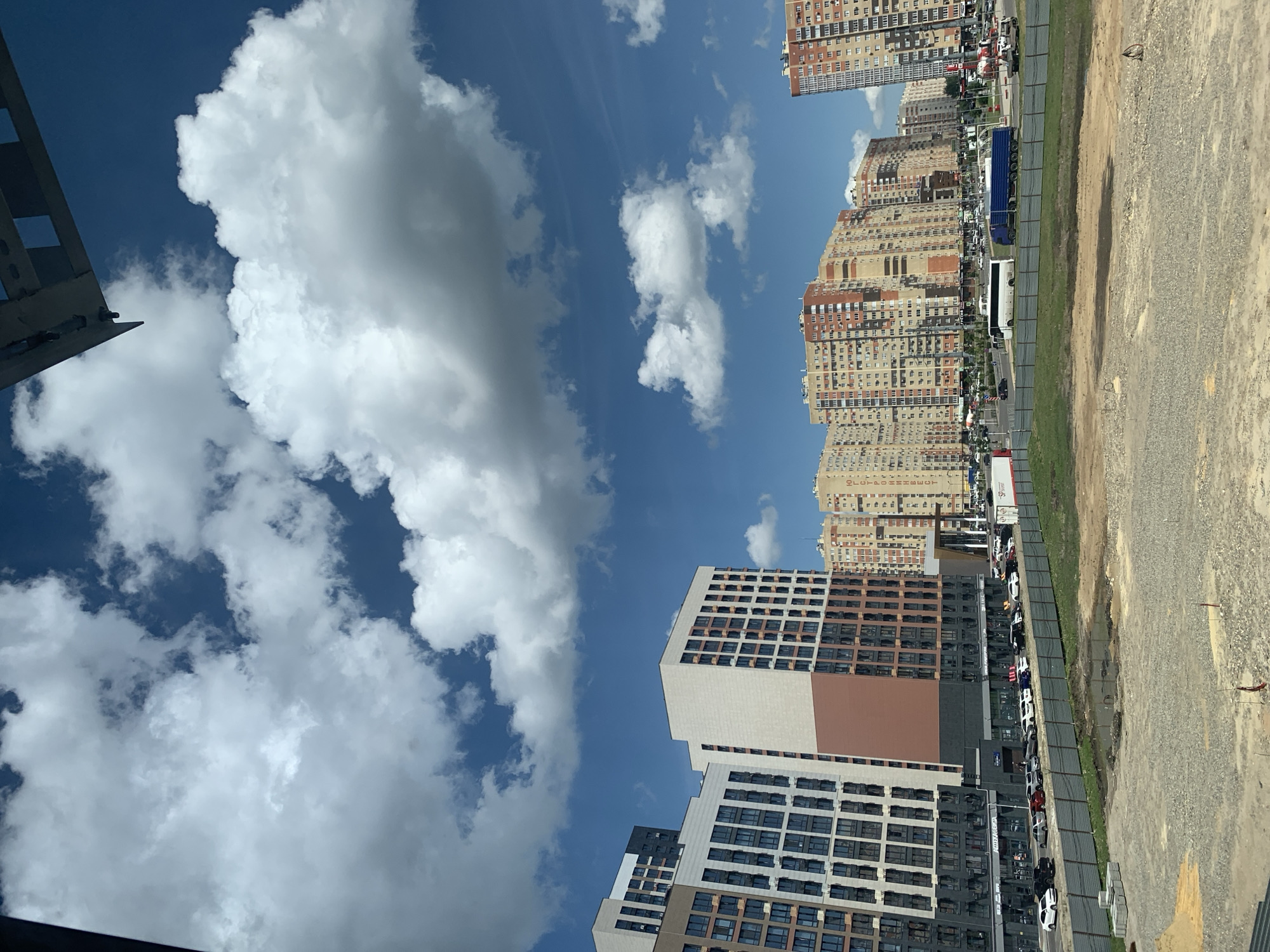 The image shows a street scene in Stavropol, Russia, on a sunny day with scattered clouds. The foreground is dominated by a view of several modern, multi-story apartment buildings. These buildings have a contemporary architectural style with many windows and a mix of beige, brown, and white facades. Some of the buildings appear to be under construction or recently completed, as evidenced by the presence of a dusty, gravel-covered lot and a temporary fence separating it from a grassy area.
Further down the street, a line of cars is parked along the roadside, and a large blue truck and a white bus are also visible. The street is lined with more apartment buildings, which are taller and have a denser appearance, suggesting a more established urban area. The sky is a clear blue, with large, fluffy white clouds drifting by. The overall impression is one of urban development and everyday life in a Russian city. There is no visible text in the image.