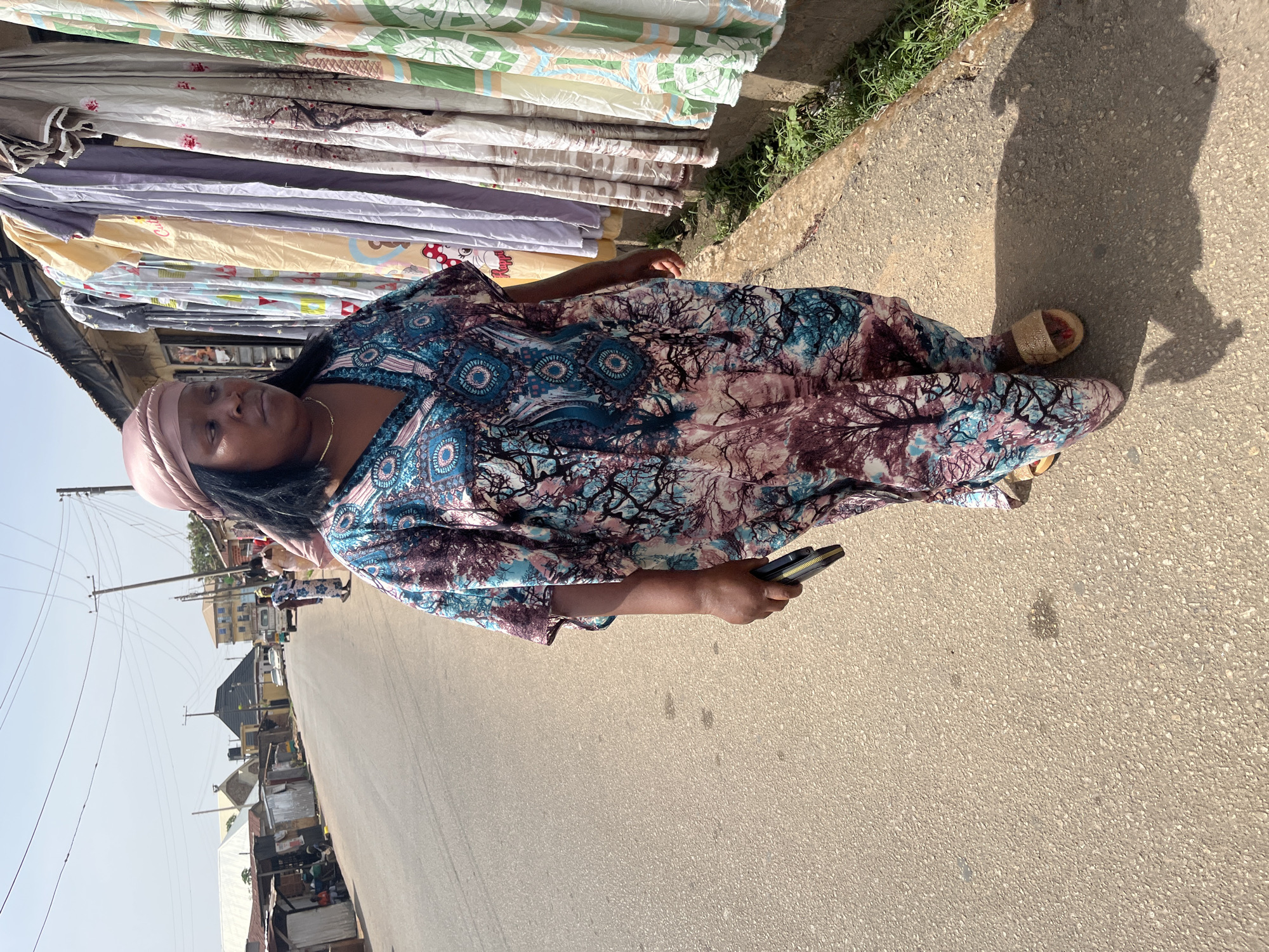 A woman stands on a paved street in Jos, Nigeria. She is dressed in a patterned dress and a headwrap. She holds a wallet in her right hand. To her left, a market stall displays folded textiles. In the background, the street stretches into the distance, revealing buildings and utility poles. Her shadow falls on the street.