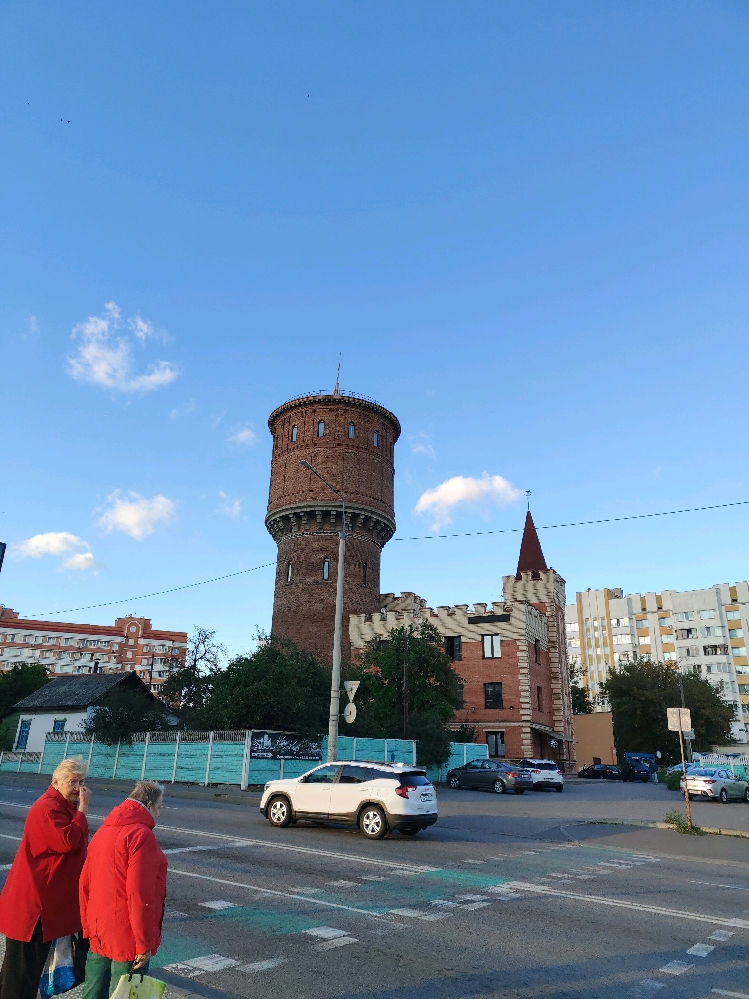 This image captures a street scene in Homyel, Belarus, on a bright, clear day with a few scattered clouds. Dominating the skyline is a tall, cylindrical brick water tower with arched windows, an architectural element reminiscent of a castle. Adjacent to the tower is a smaller brick building with crenellated battlements and a pointed, red-tiled roof, also evoking a castle-like aesthetic.
In the foreground, two older women are walking near a pedestrian crossing. They are both wearing red coats, one a longer, flowing style and the other a more structured jacket. The woman on the left appears to be touching her face or hair, while the woman on the right is looking forward. Both are carrying bags.
The street is paved, with a clear pedestrian crossing marked by white lines and green accents. Several cars are visible on the road and in a parking area behind a turquoise fence. These include a white SUV, several sedans, and a darker vehicle. In the background, residential buildings with multiple floors line the street, some with balconies and apartment windows. Trees and foliage are interspersed throughout the scene, suggesting a moderately urban environment.
The lighting indicates it is likely late afternoon or early evening, given the warm tones and the long shadows cast. The sky is a vibrant blue, and the overall atmosphere appears calm and ordinary, depicting a moment of everyday life in the city. There is no visible text in the image.