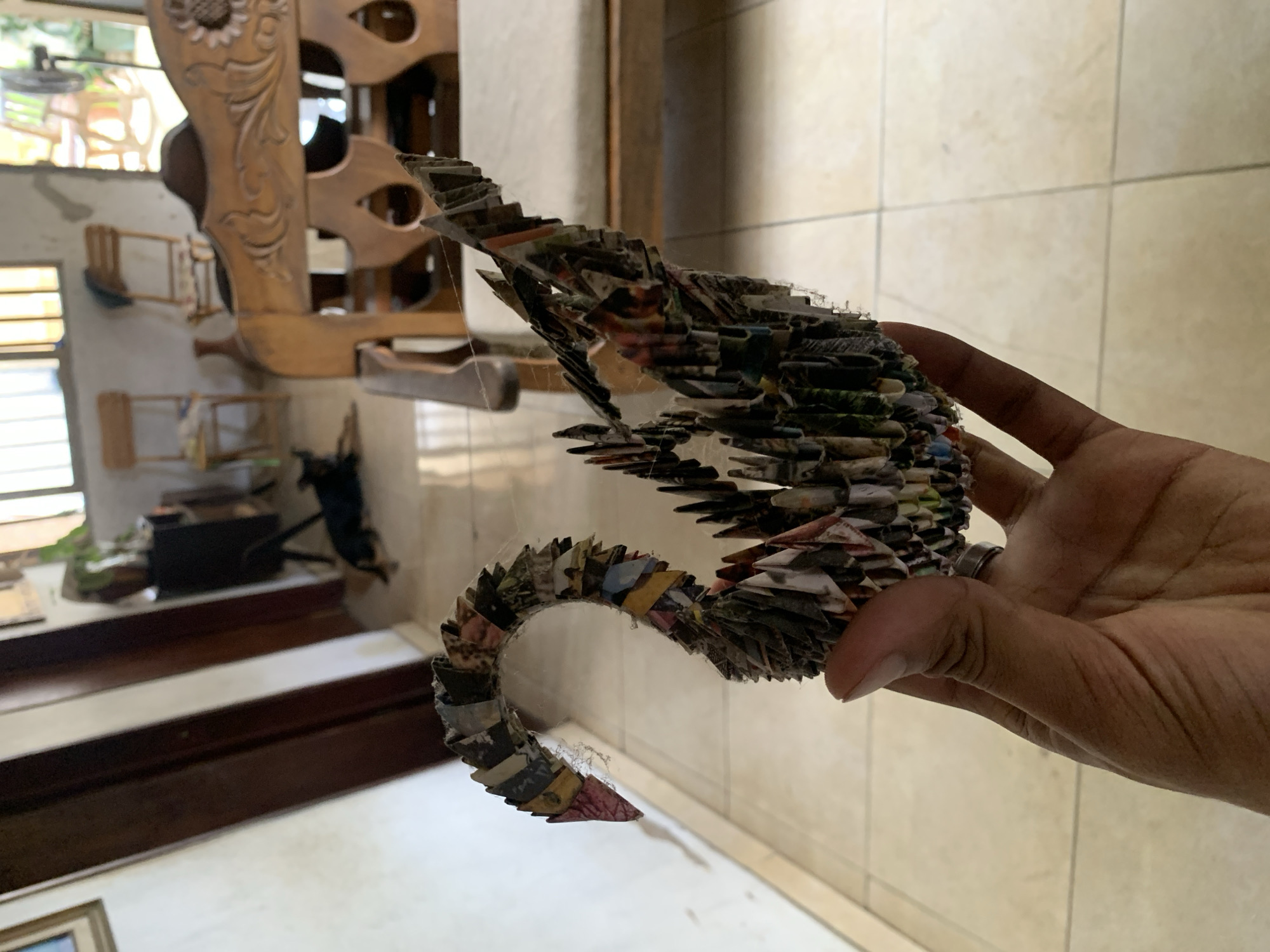 A hand holds a sculpture made of rolled-up paper in front of a tiled wall. The sculpture resembles a stylized creature with a curved tail and a ridged back. In the background, there is a carved wooden structure and glimpses of a room with various objects. The location is Santa Ana, Venezuela.