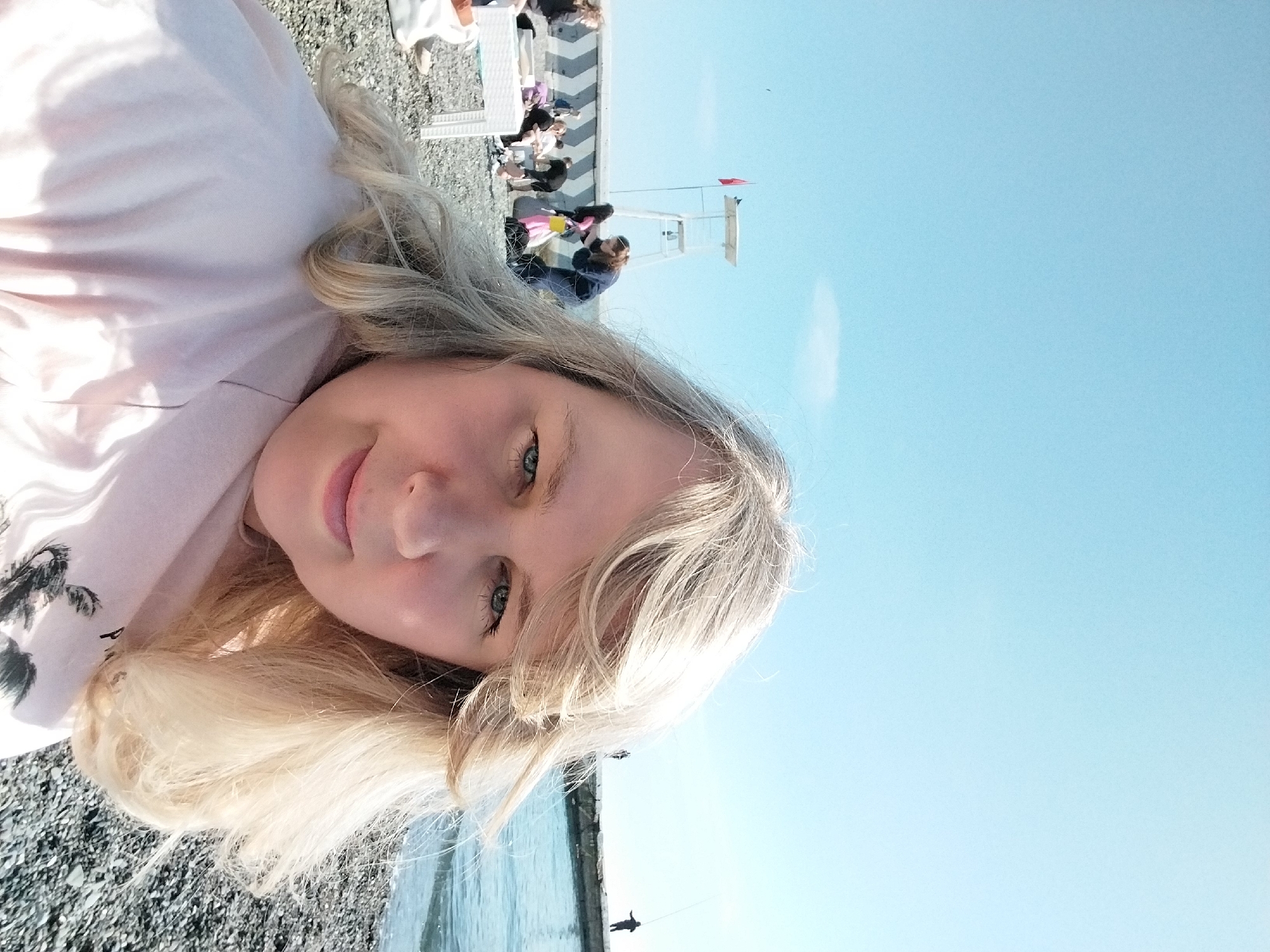 A young woman with long, wavy blonde hair and blue eyes is prominently featured in the foreground of this outdoor photograph, taking a selfie. She has a slight, gentle smile on her face and is looking directly at the camera. She is wearing a light pink or peach-colored long-sleeved top, possibly a hoodie, with a dark graphic design, resembling palm trees, visible on her left shoulder.
The setting is a sunny beach or coastal area, suggested to be in Sochi, Russia. The ground in the foreground is covered with dark pebbles or small stones. To the right of the woman and extending into the mid-ground is a body of calm, blue water, likely the sea or ocean.
In the mid-distance, behind the woman and further along the beach, several other people are visible. Some are seated in white beach chairs, while others stand near a white lifeguard stand that has a small red flag at its top. There's also a paved or patterned surface visible near these people, suggesting a promenade or organized beach area. In the far distance, to the right, a solitary figure can be seen fishing from what appears to be a pier or breakwater that extends into the water.
The sky, dominating the upper right portion of the image, is a clear and bright blue, indicating sunny weather during what appears to be mid-morning or afternoon. The overall atmosphere of the scene is relaxed and pleasant. No discernible text is visible in the image.